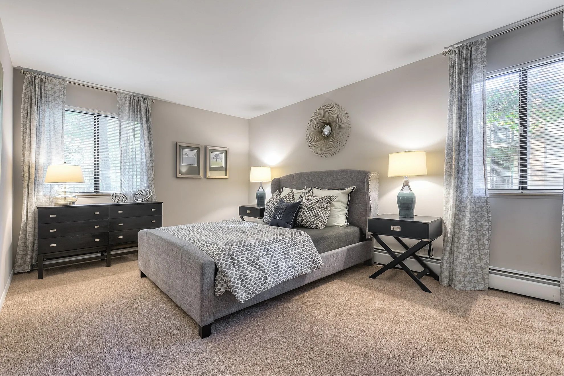 Chic and cozy bedroom with a plush grey bed, stylish decor, and ample natural light through large windows.