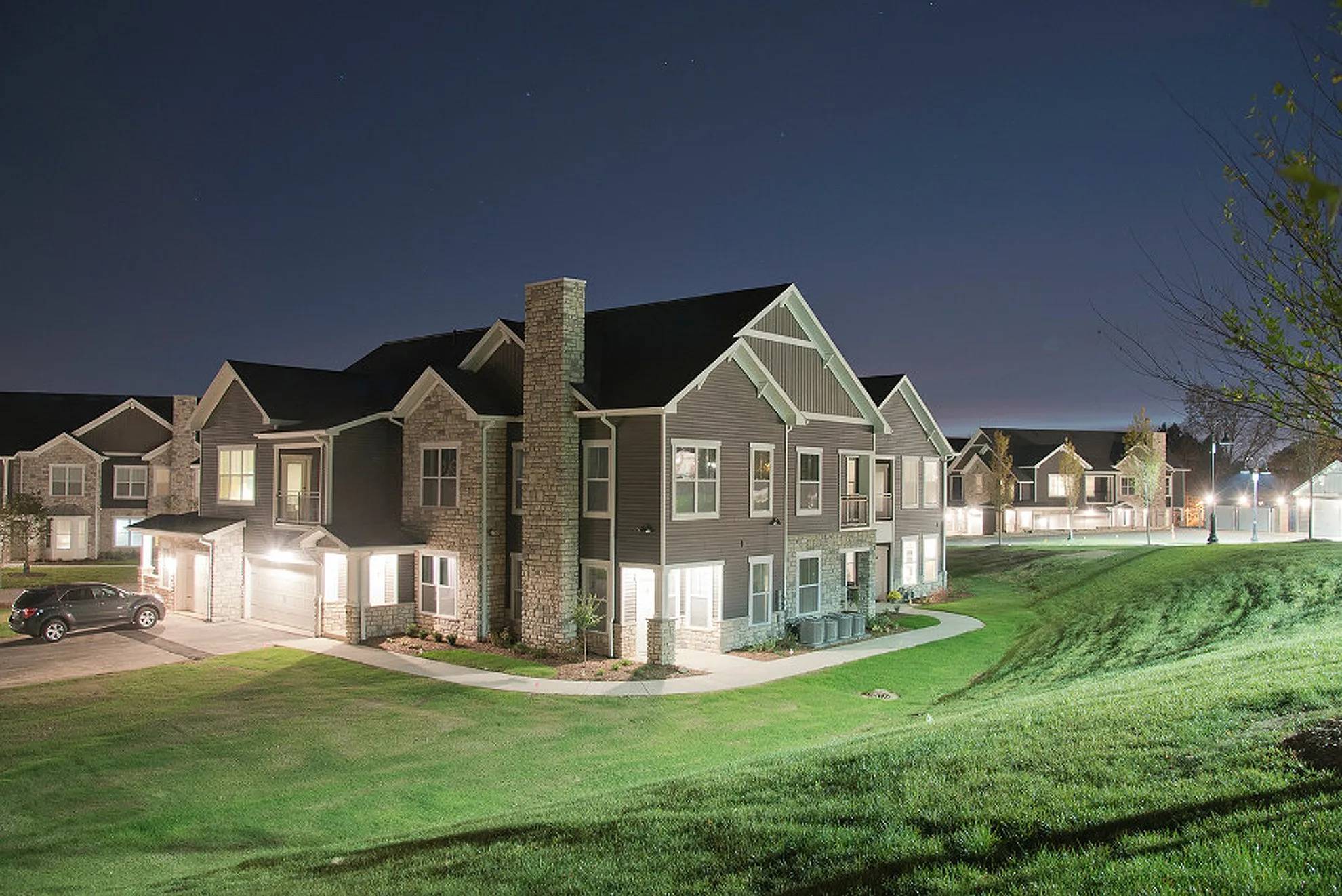 Charming two-story apartment complex with stone accents, beautifully lit against a serene night sky, surrounded by lush green lawns.