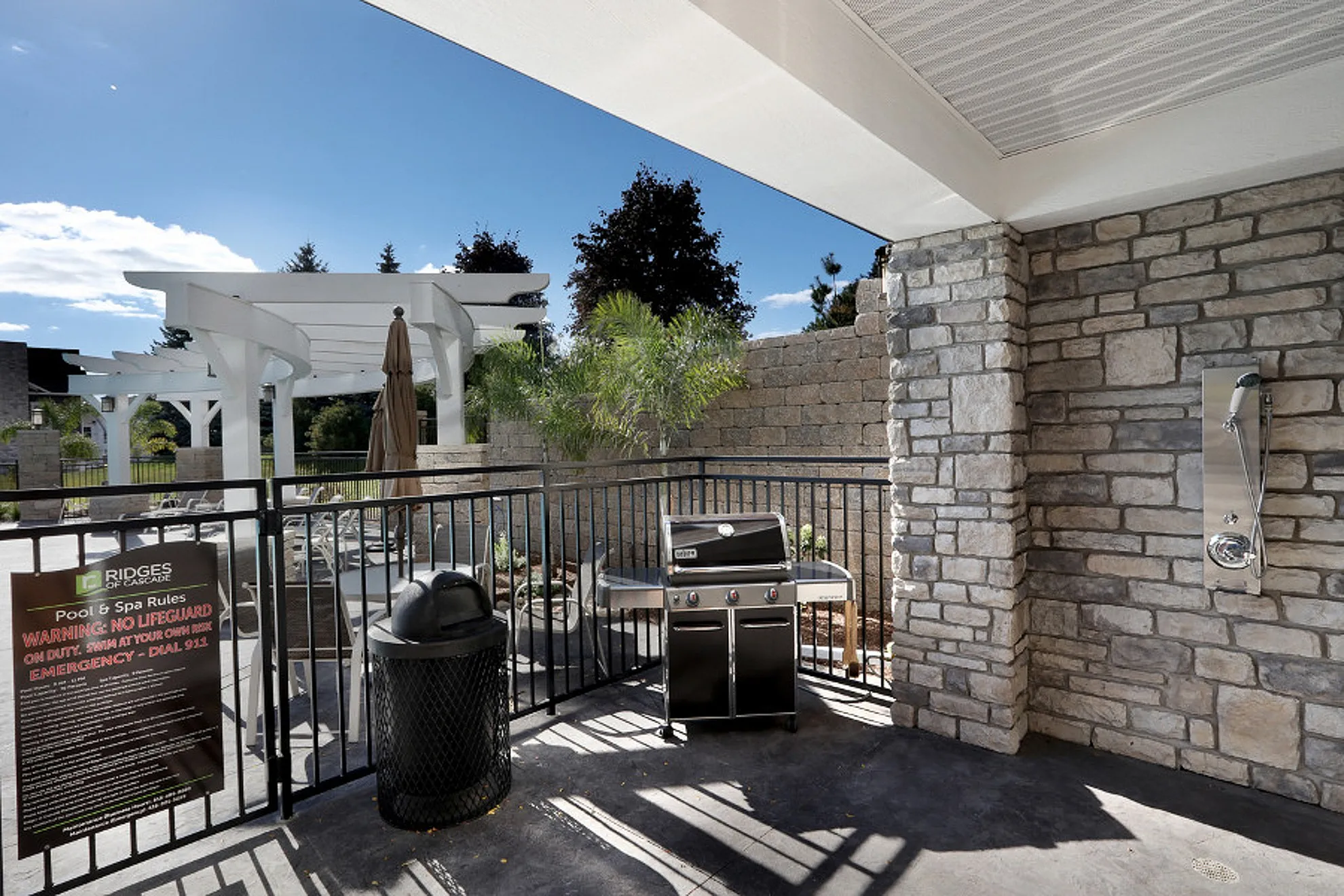 Charming outdoor area with a modern grill and elegant stone walls, complemented by lush greenery and a stylish pergola under a bright blue sky.