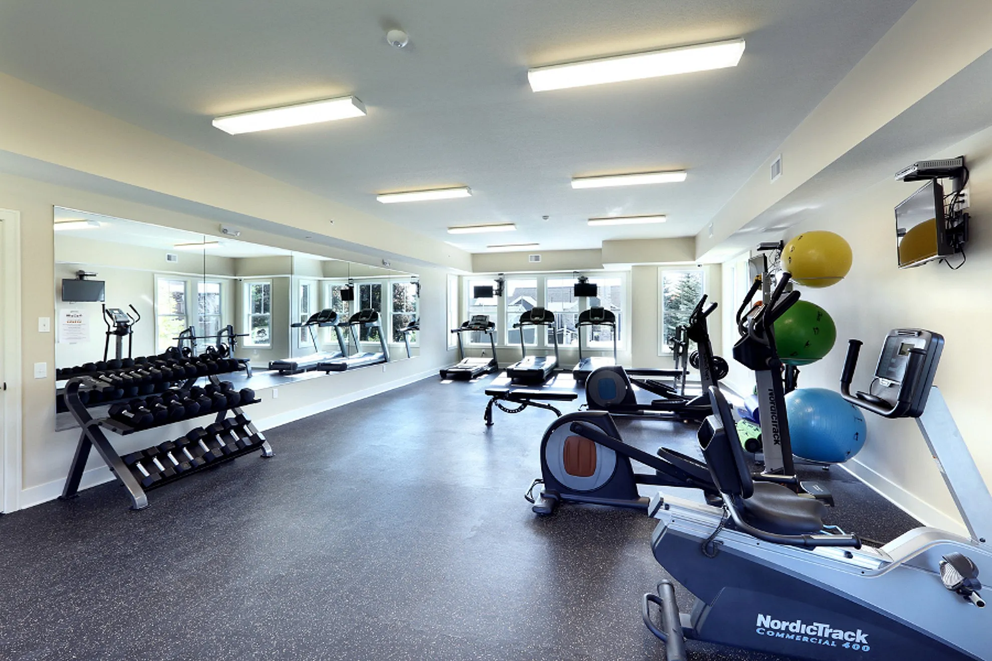 Modern fitness center featuring state-of-the-art treadmills, a weight rack, and exercise bikes, with large mirrors and bright windows creating a spacious and inviting atmosphere.
