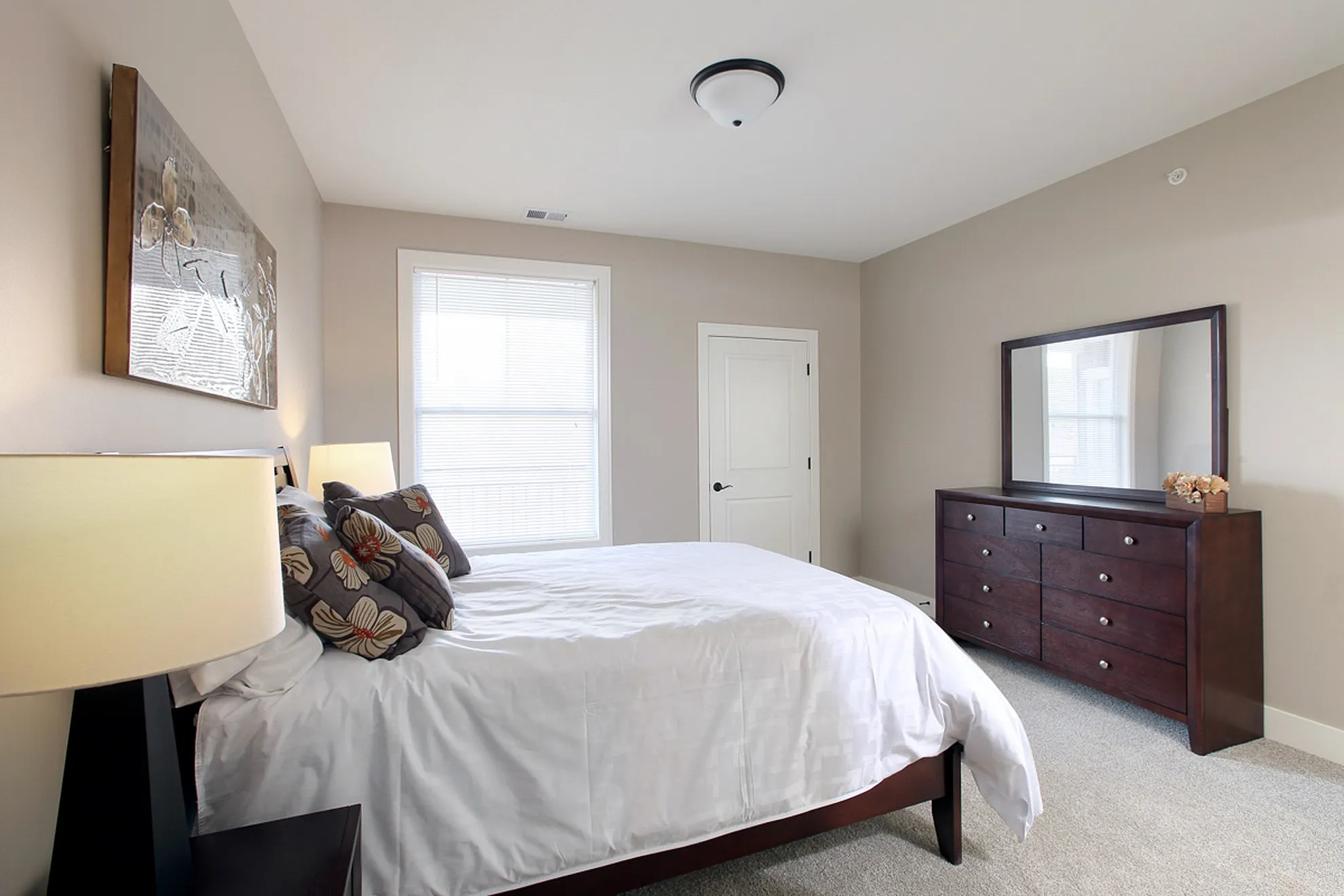 Bright and cozy bedroom with elegant decor, featuring a plush bed with floral pillows, a stylish dark wood dresser, and a large window inviting natural light.