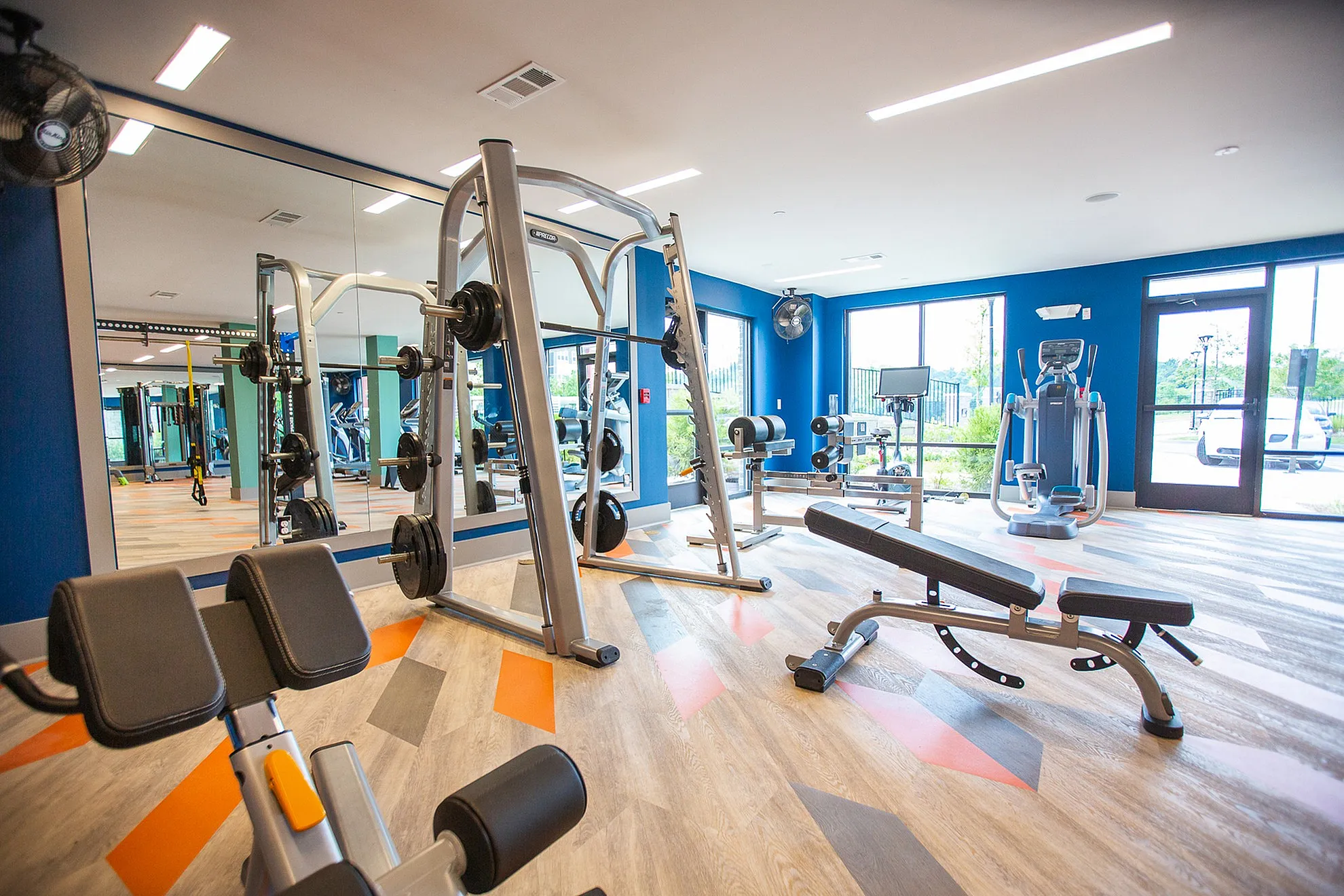 Modern gym room with high-end fitness equipment and vibrant design, featuring large windows and stylish flooring.