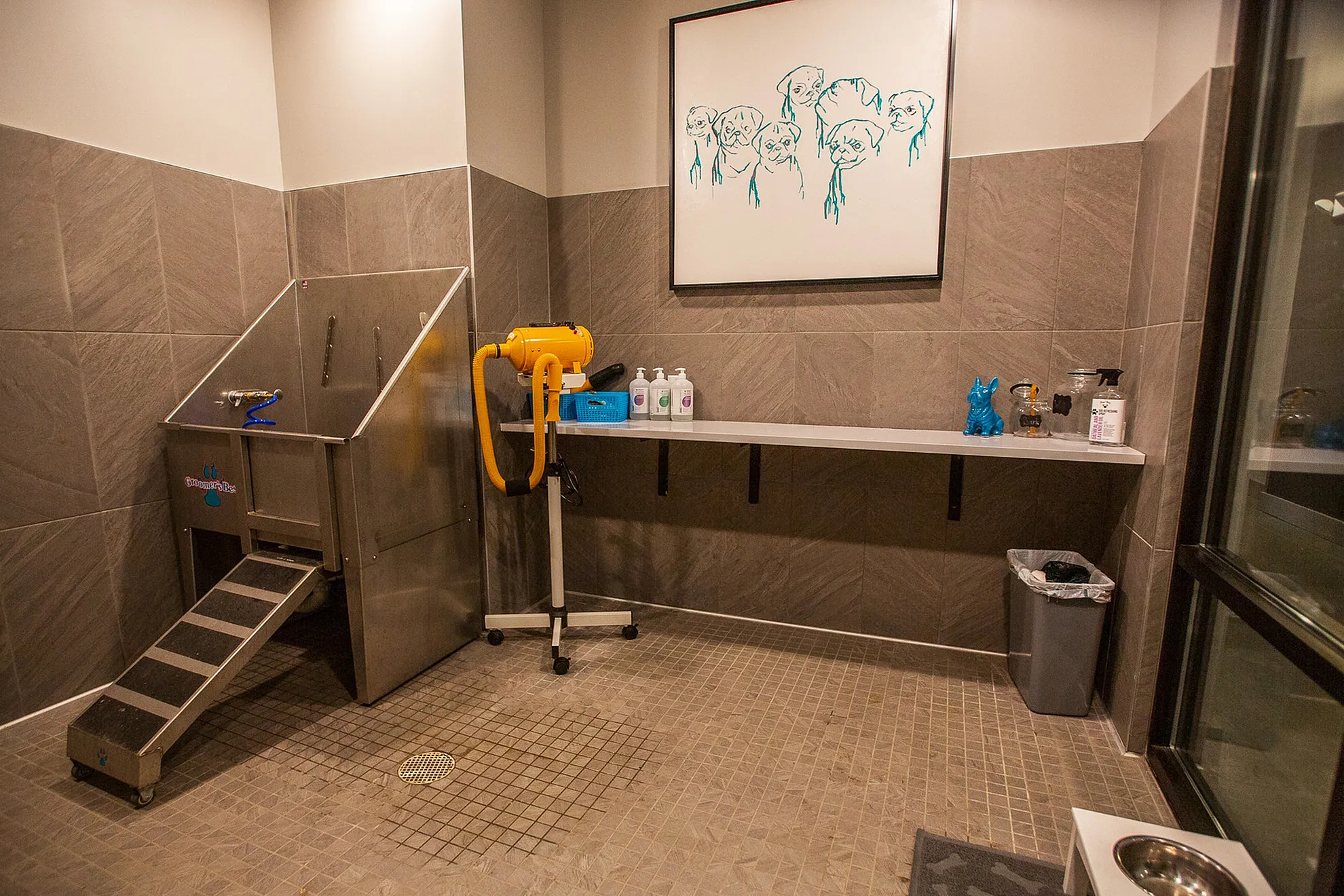 Modern pet grooming room with stainless steel washing station, artistic dog portrait, and stylish tiled design.