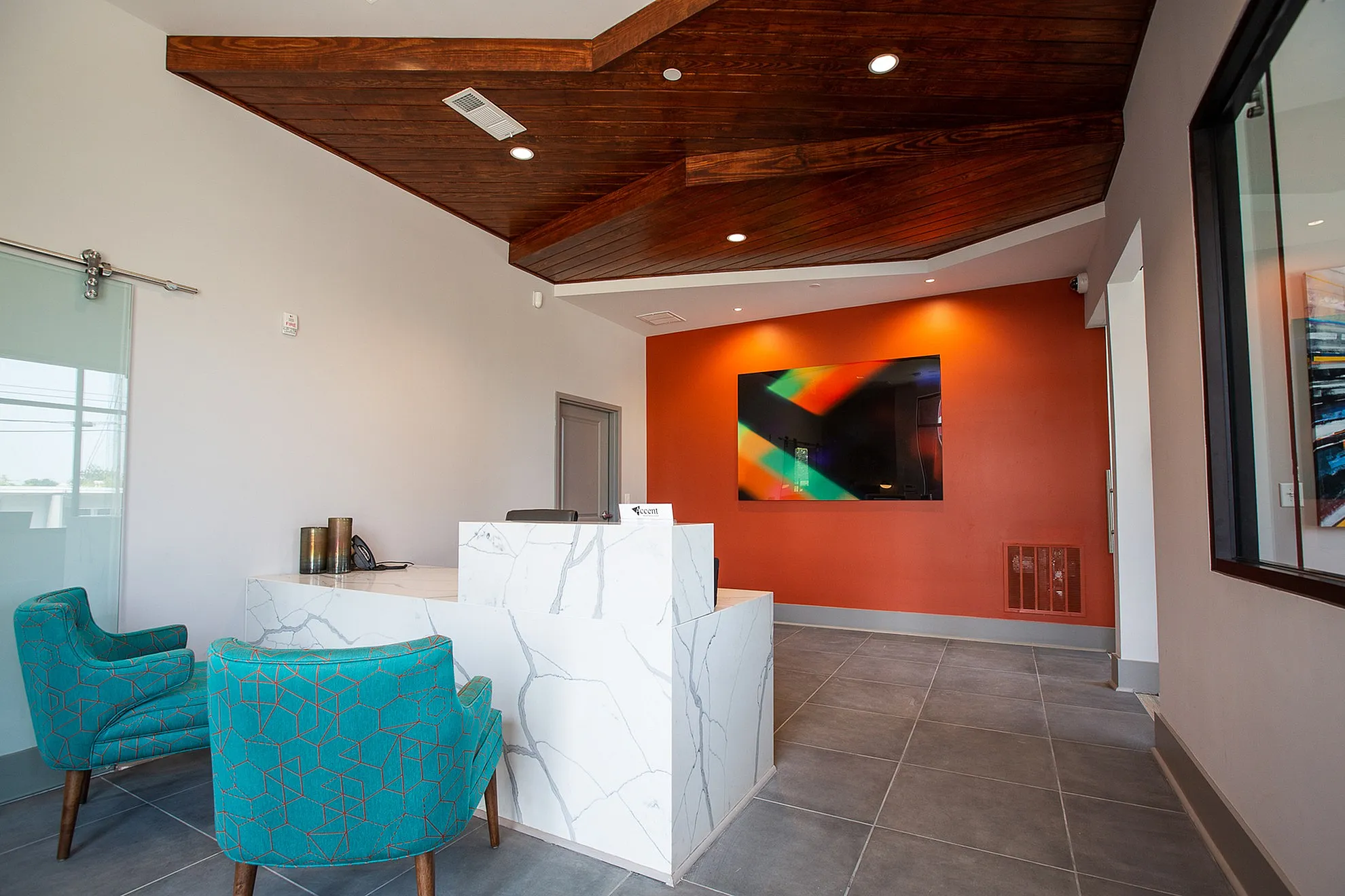 Contemporary reception area with vibrant orange accent wall, marble desk, and stylish teal chairs under a vaulted wooden ceiling.