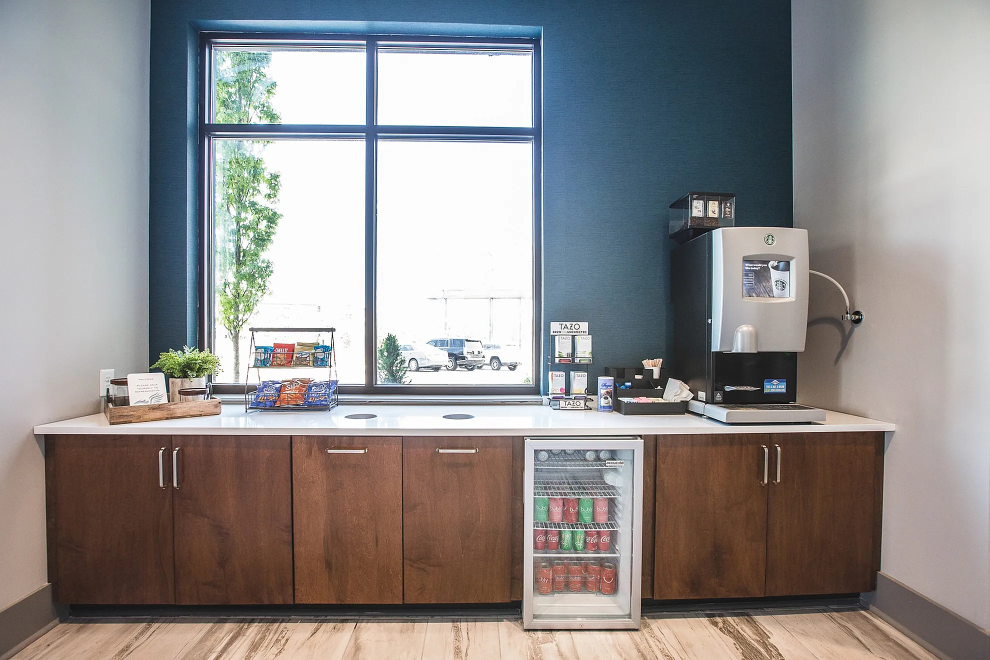 Chic refreshment corner with a sleek coffee machine, snacks, and a mini fridge stocked with beverages, set against a stylish blue wall and elegant wooden cabinetry.