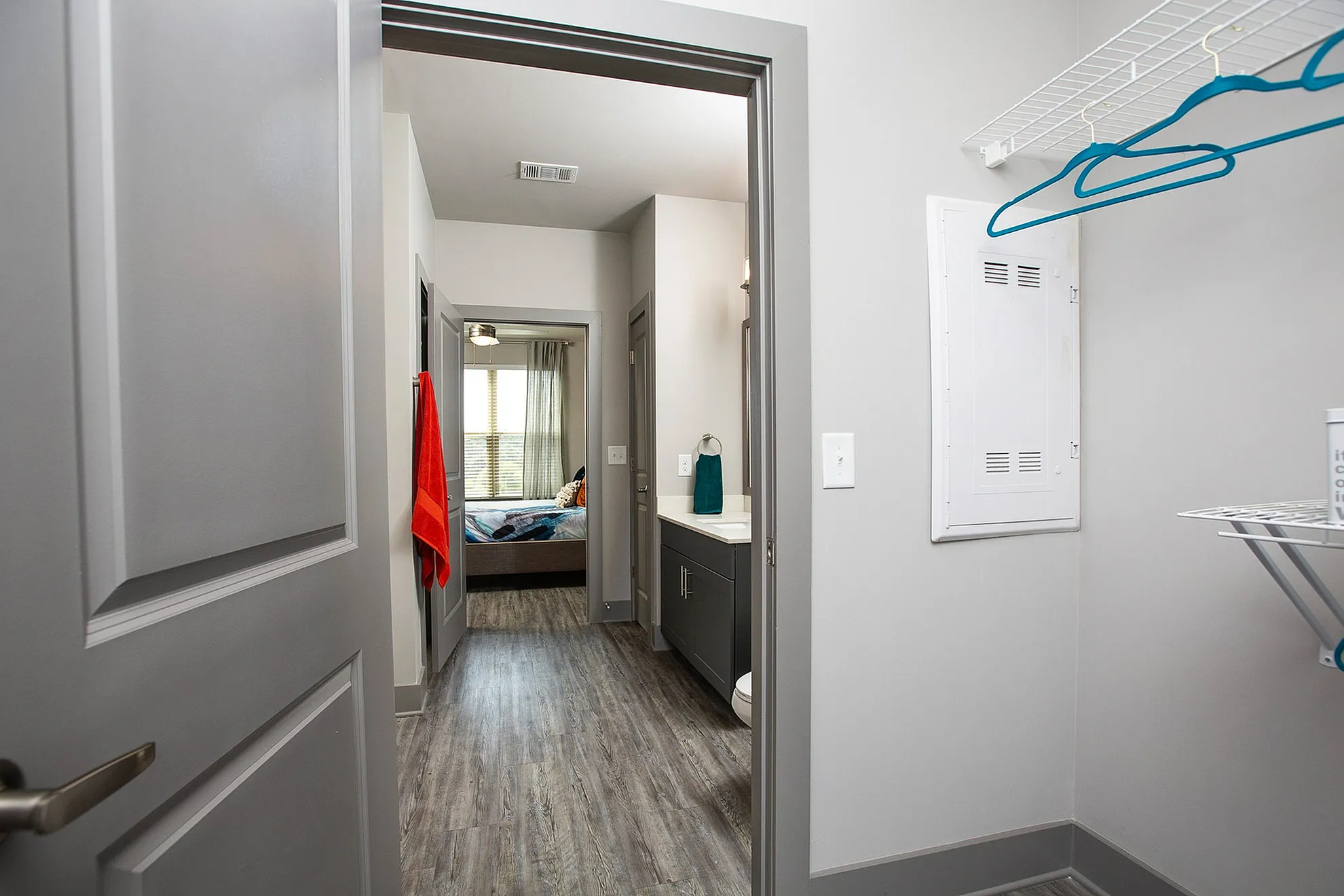 Modern apartment hallway with stylish wood flooring leading to a well-lit bedroom, featuring a sleek bathroom vanity and vibrant towel accents.