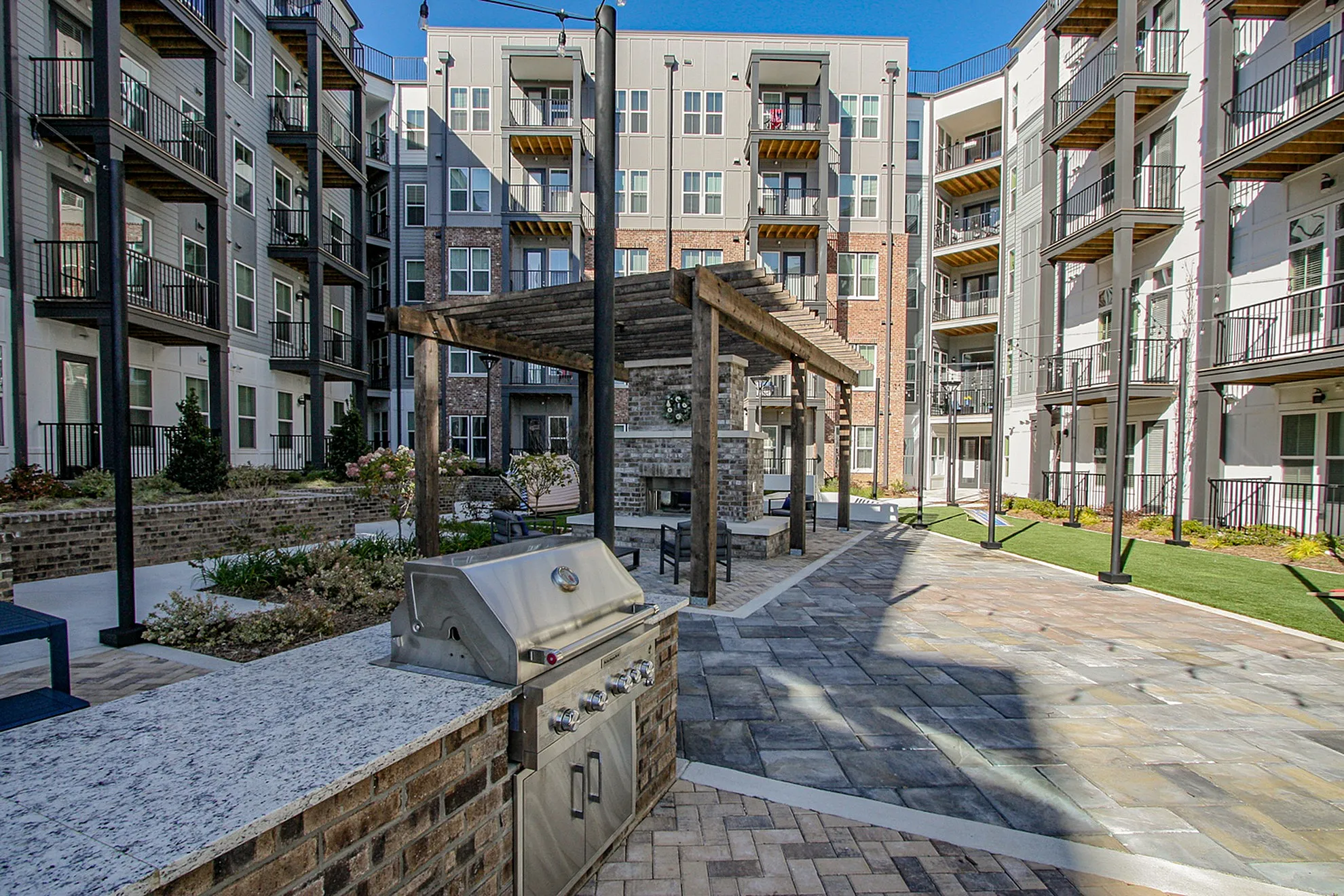Modern apartment complex courtyard featuring a stylish pergola, outdoor seating, and a sleek grill station, surrounded by lush landscaping and contemporary balconies.