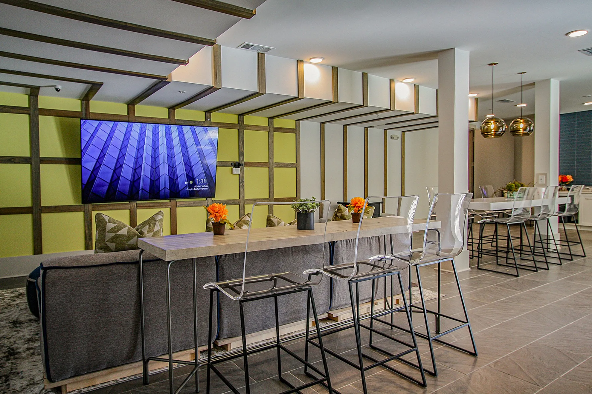 Modern communal area featuring a sleek TV wall, stylish bar seating with transparent chairs, and vibrant floral decor.