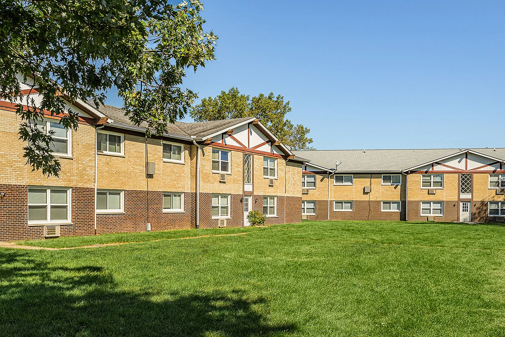 Charming apartment building with classic brick facade, surrounded by lush green lawn and shaded by leafy trees on a sunny day.