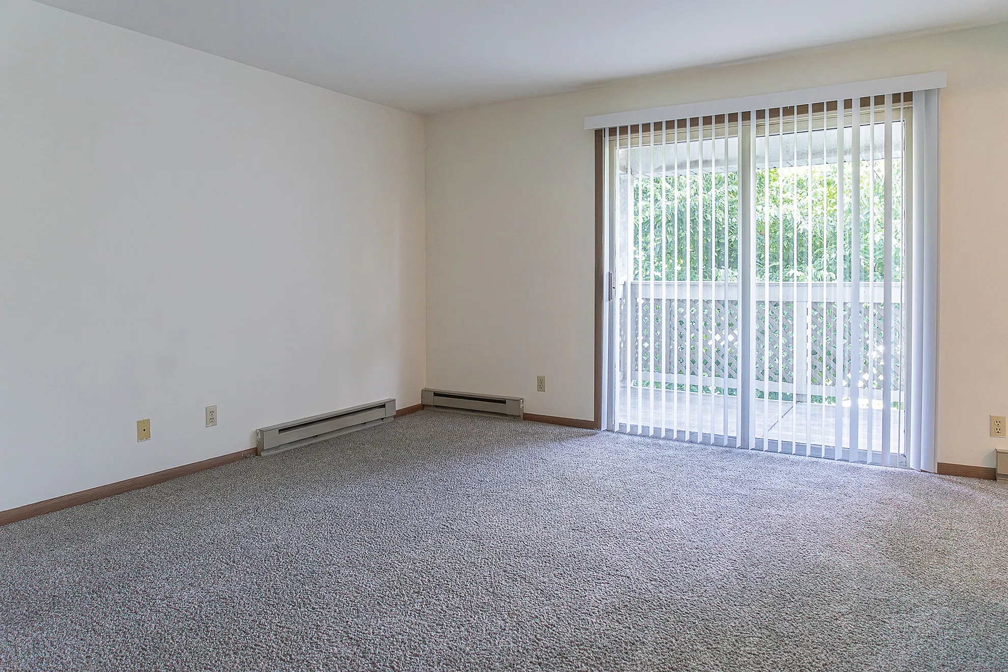 Bright and airy living space with plush carpet, neutral walls, and large sliding glass doors opening to a private balcony with lush greenery view.