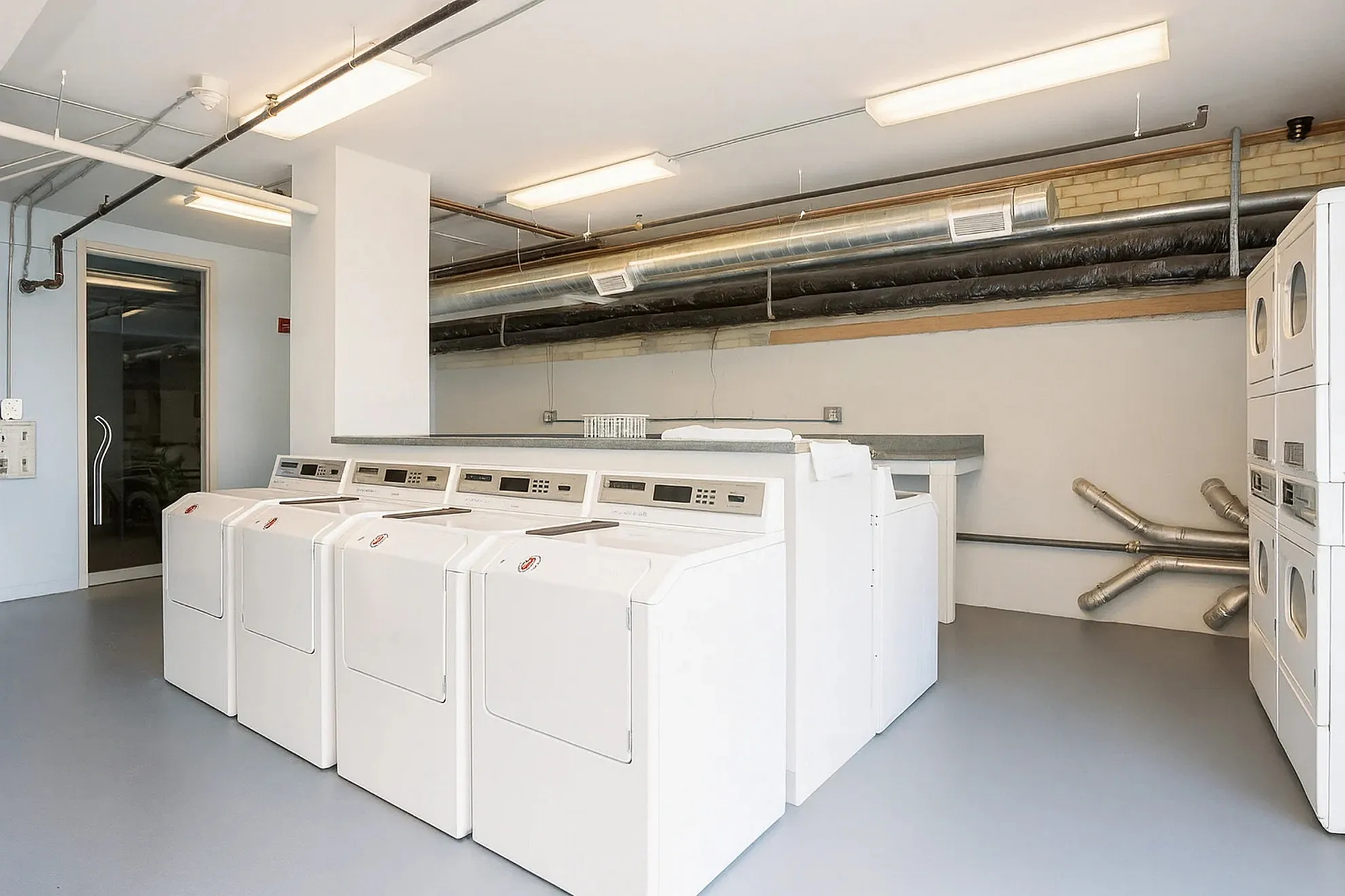 Bright and modern laundry room featuring multiple washers and dryers, with sleek industrial piping adding a contemporary touch.