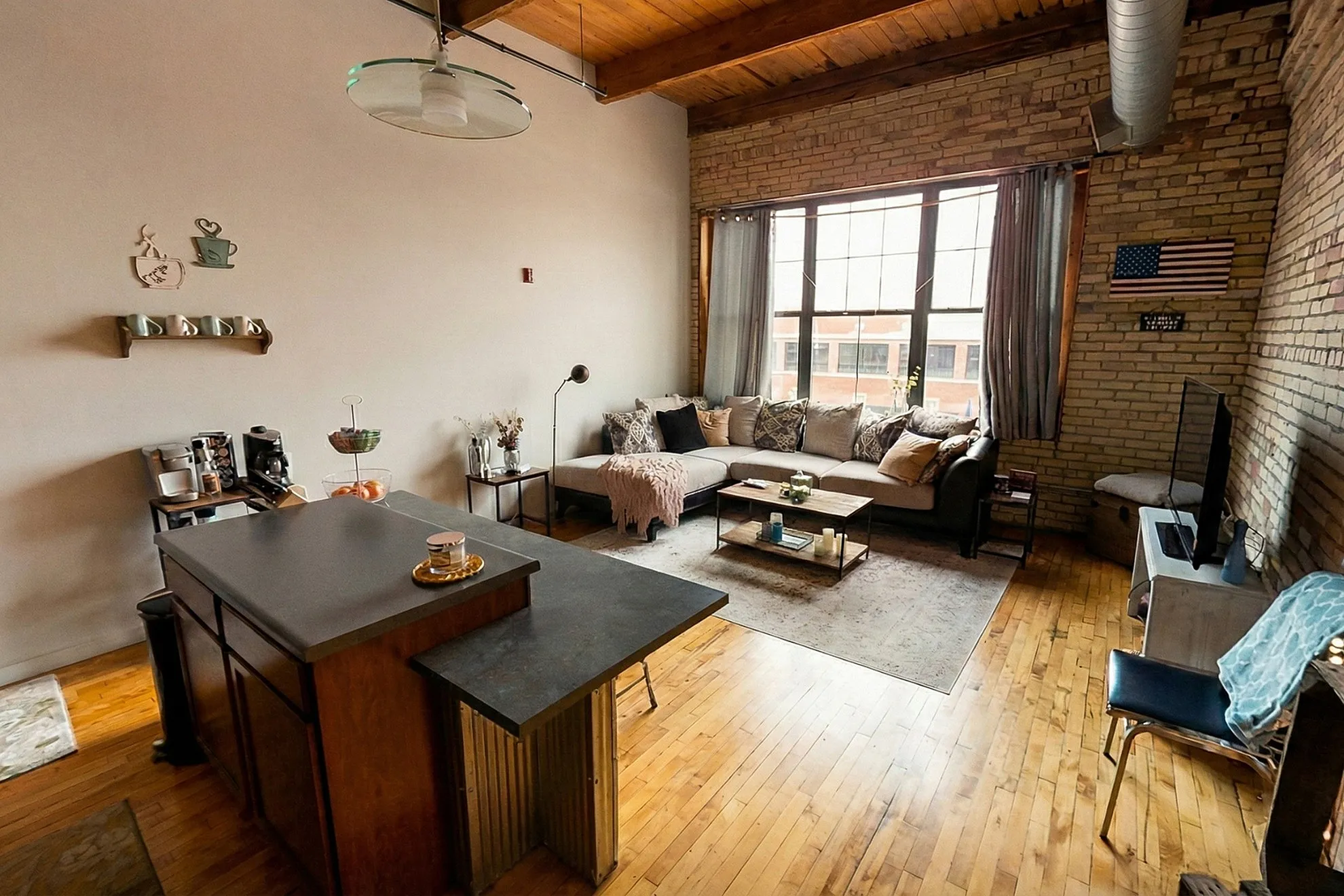 Charming loft-style living room with exposed brick, warm wooden flooring, and abundant natural light. Cozy sectional sofa, modern kitchen island, and stylish decor create an inviting atmosphere.