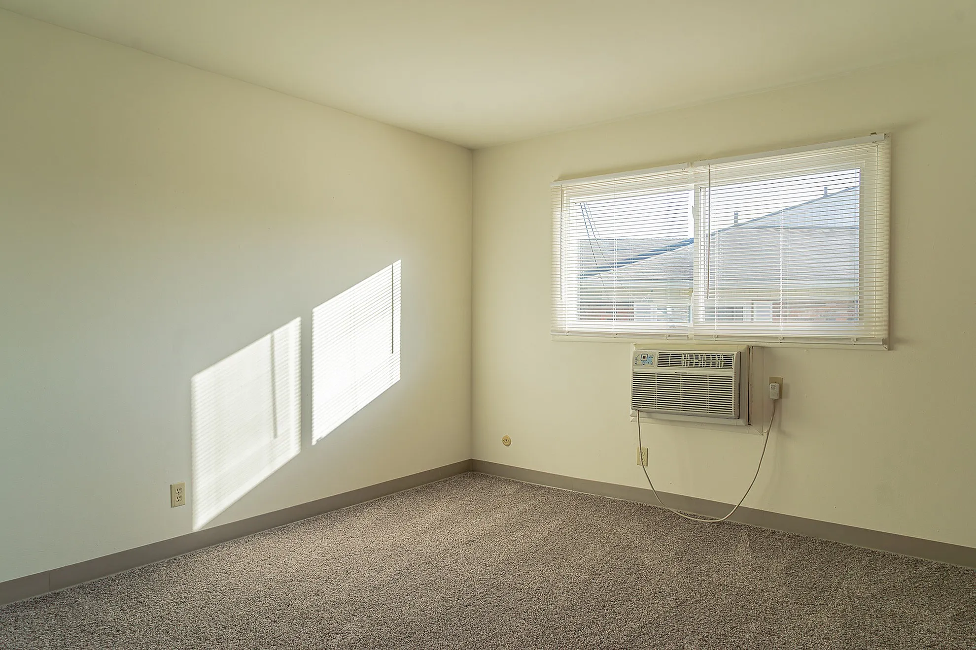Bright and cozy room featuring soft carpet flooring, large window with blinds, and a compact air conditioning unit.