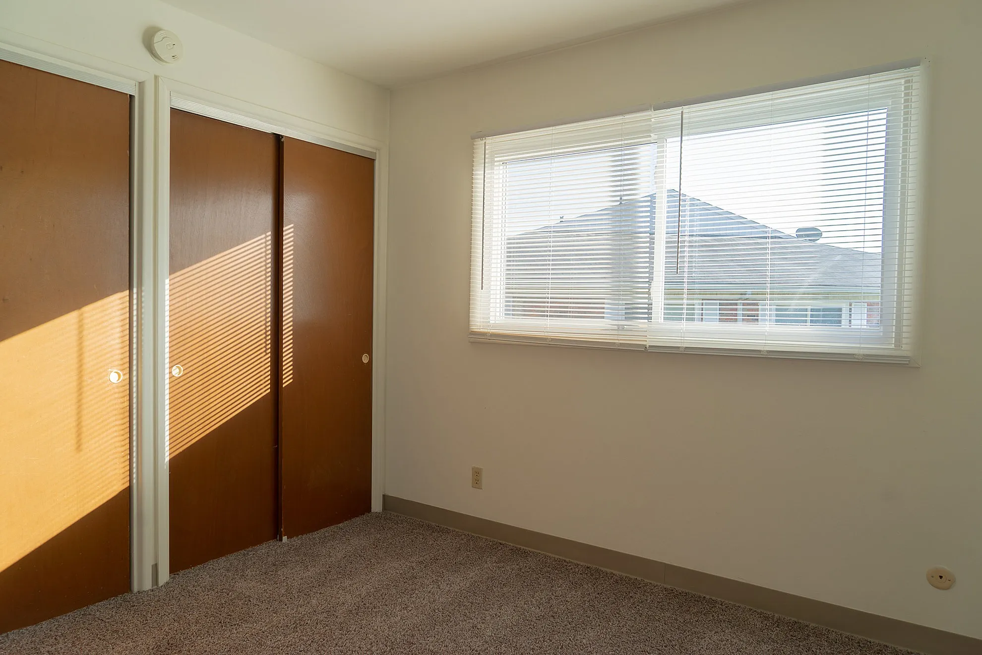 Bright bedroom featuring large window with blinds, warm wooden closet doors, and soft carpet flooring.