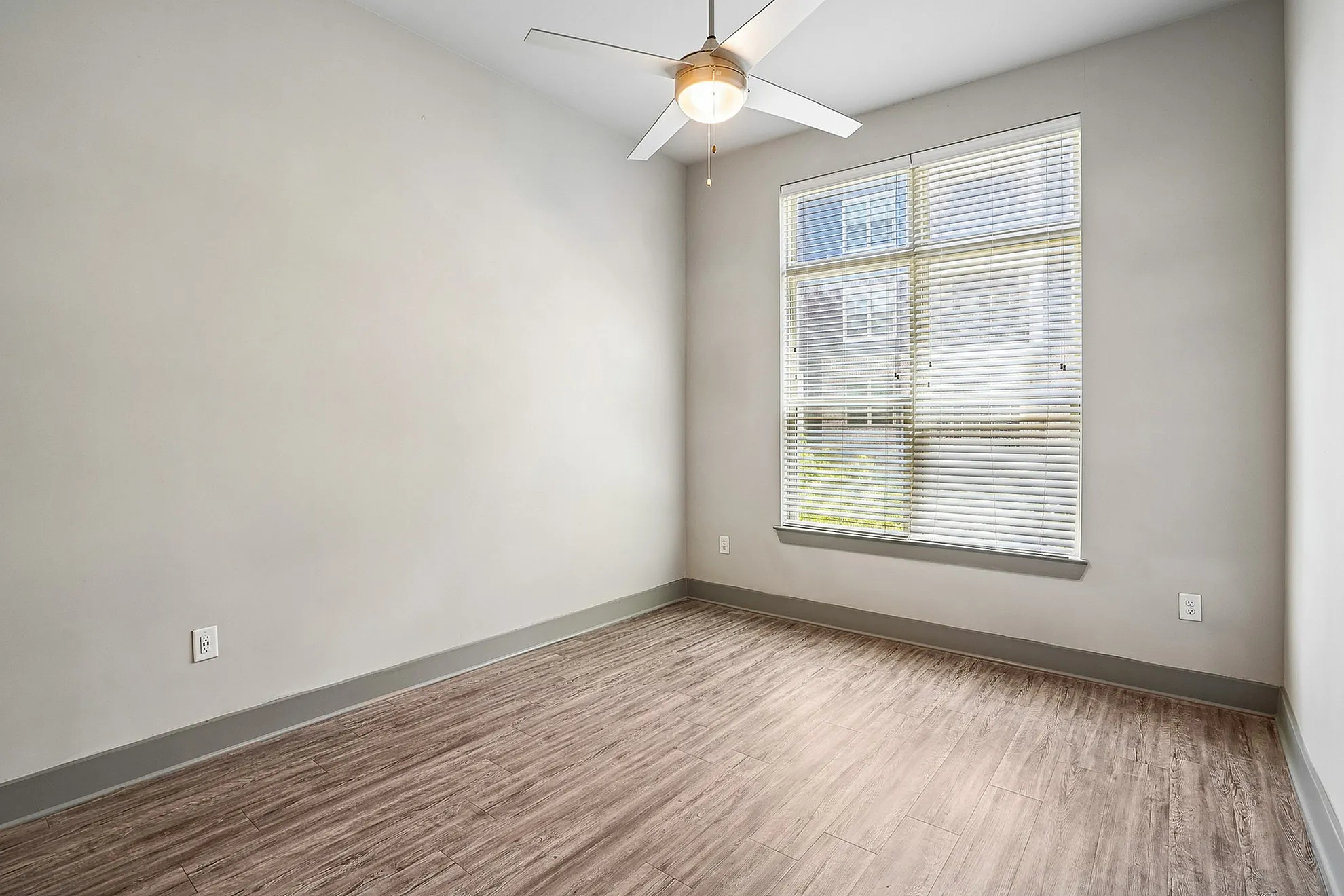 Bright and airy unfurnished room with elegant wooden floors, a large window with blinds, and a modern ceiling fan.