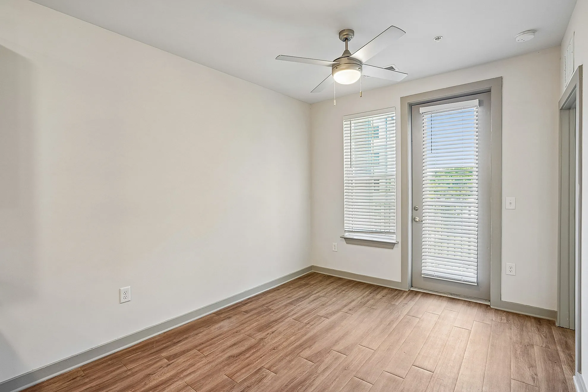 Bright apartment room with elegant wooden flooring, a modern ceiling fan, and abundant natural light through large windows with blinds.