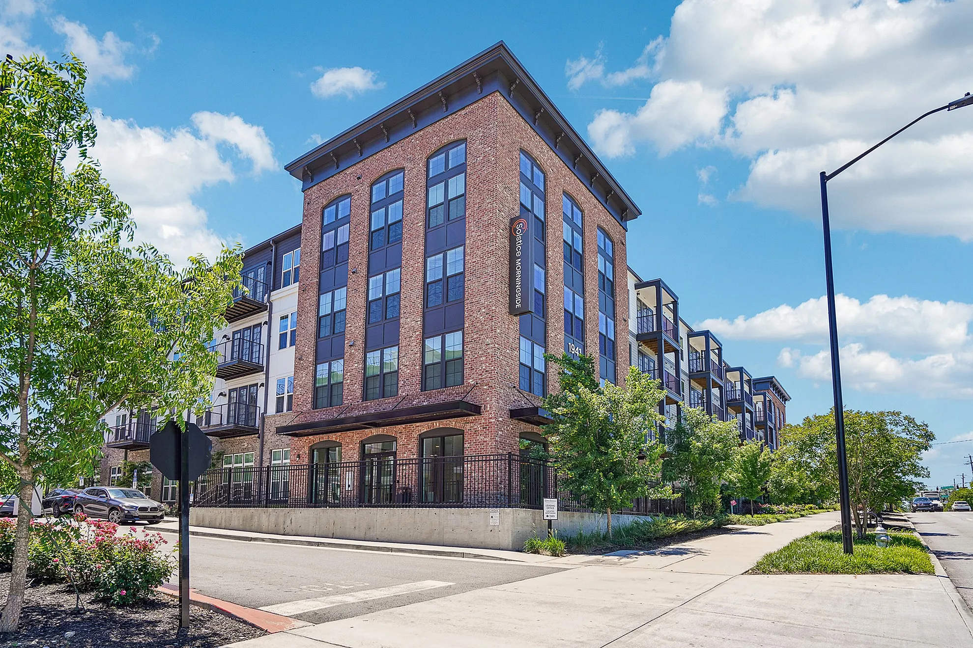 Contemporary multi-story brick apartment building with large windows, surrounded by lush greenery on a sunny day.