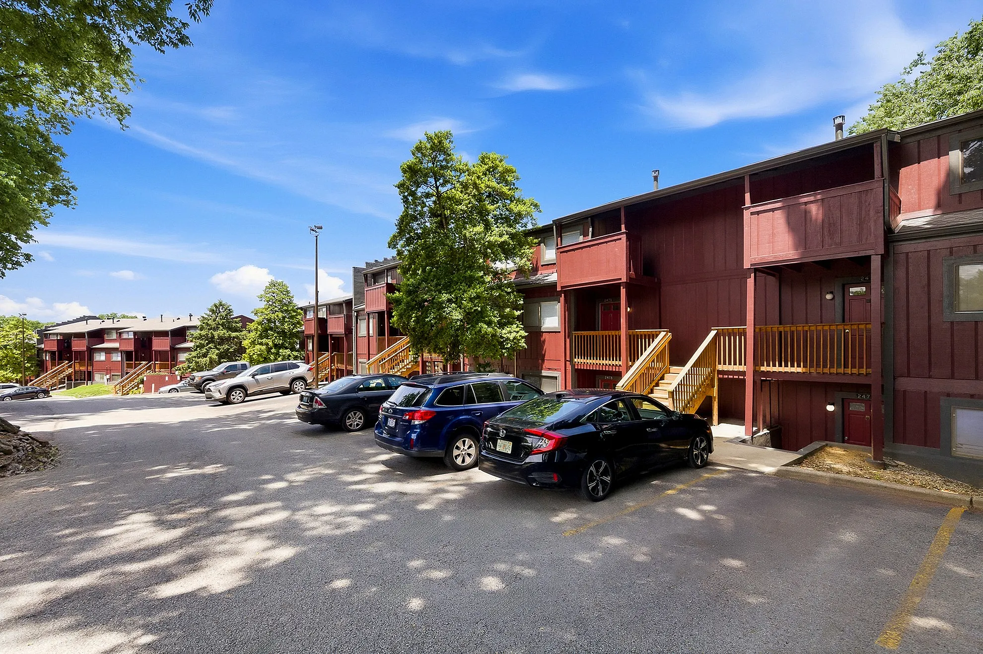 Inviting apartment complex with rich red façades and charming wooden balconies, nestled among lush greenery under a vibrant blue sky.