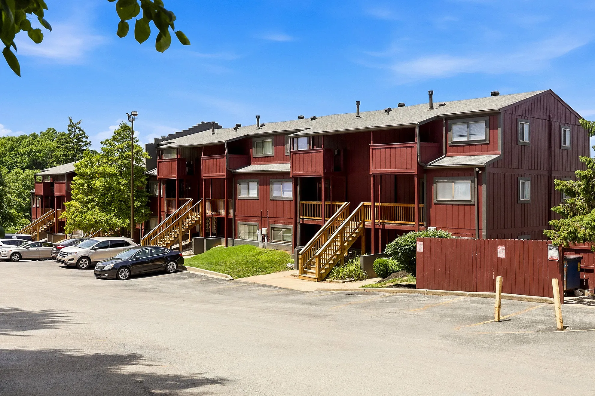 Charming three-story apartment complex with rustic red exterior, surrounded by lush greenery and convenient parking.