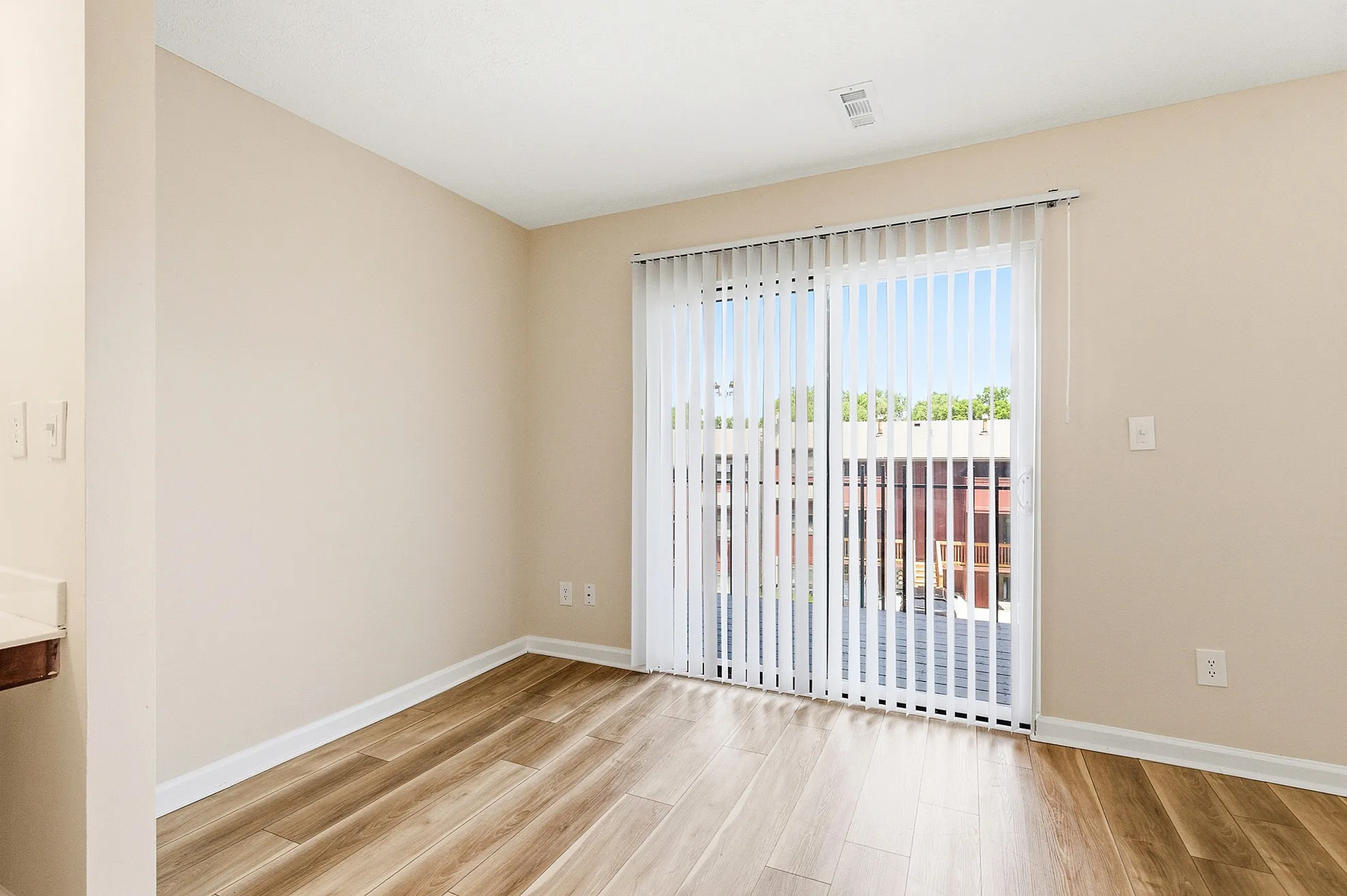 Bright and inviting living space with sleek hardwood flooring and large windows with vertical blinds, offering abundant natural light.