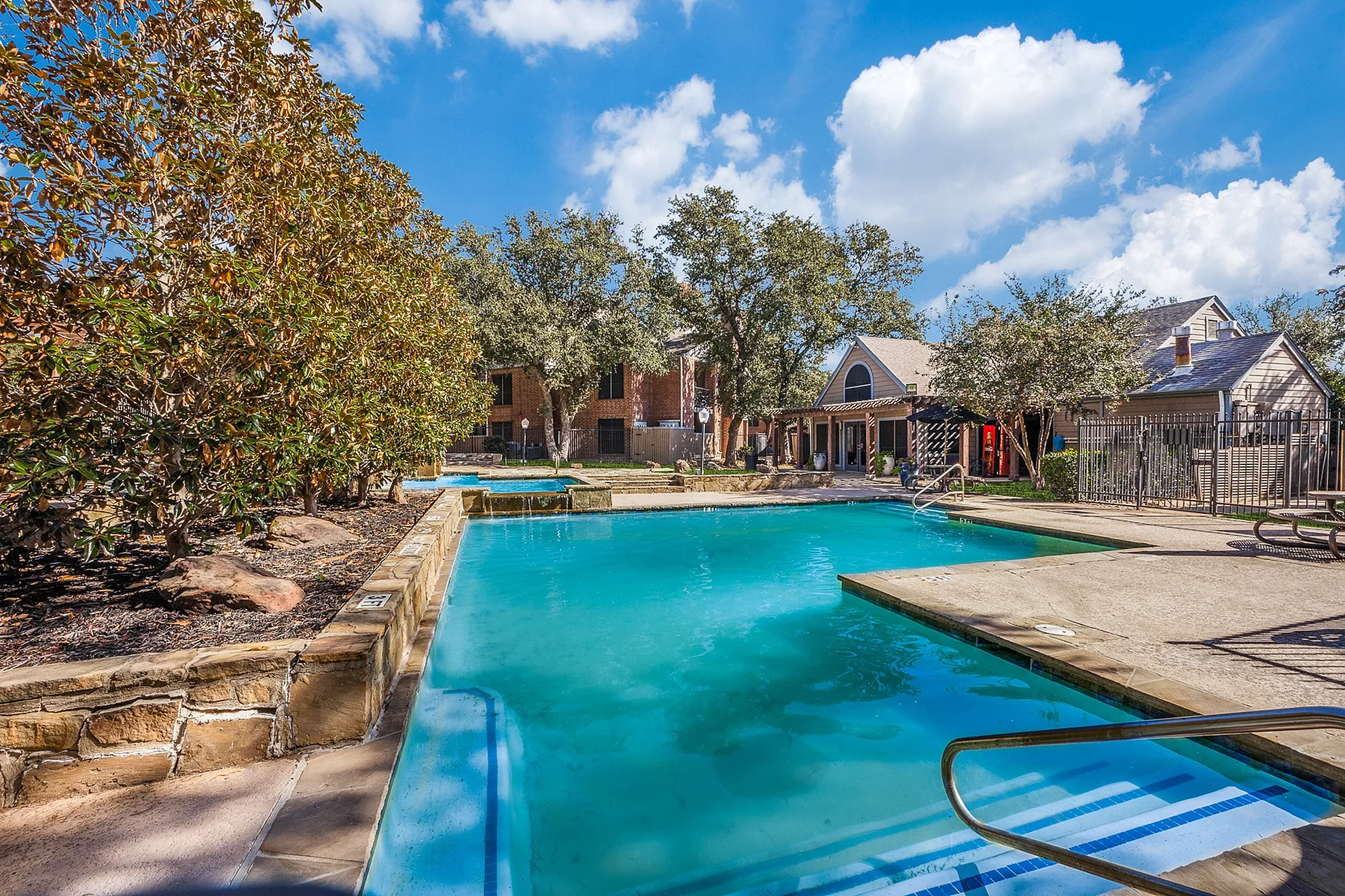 Inviting blue swimming pool surrounded by lush greenery and charming brick buildings under a bright blue sky.