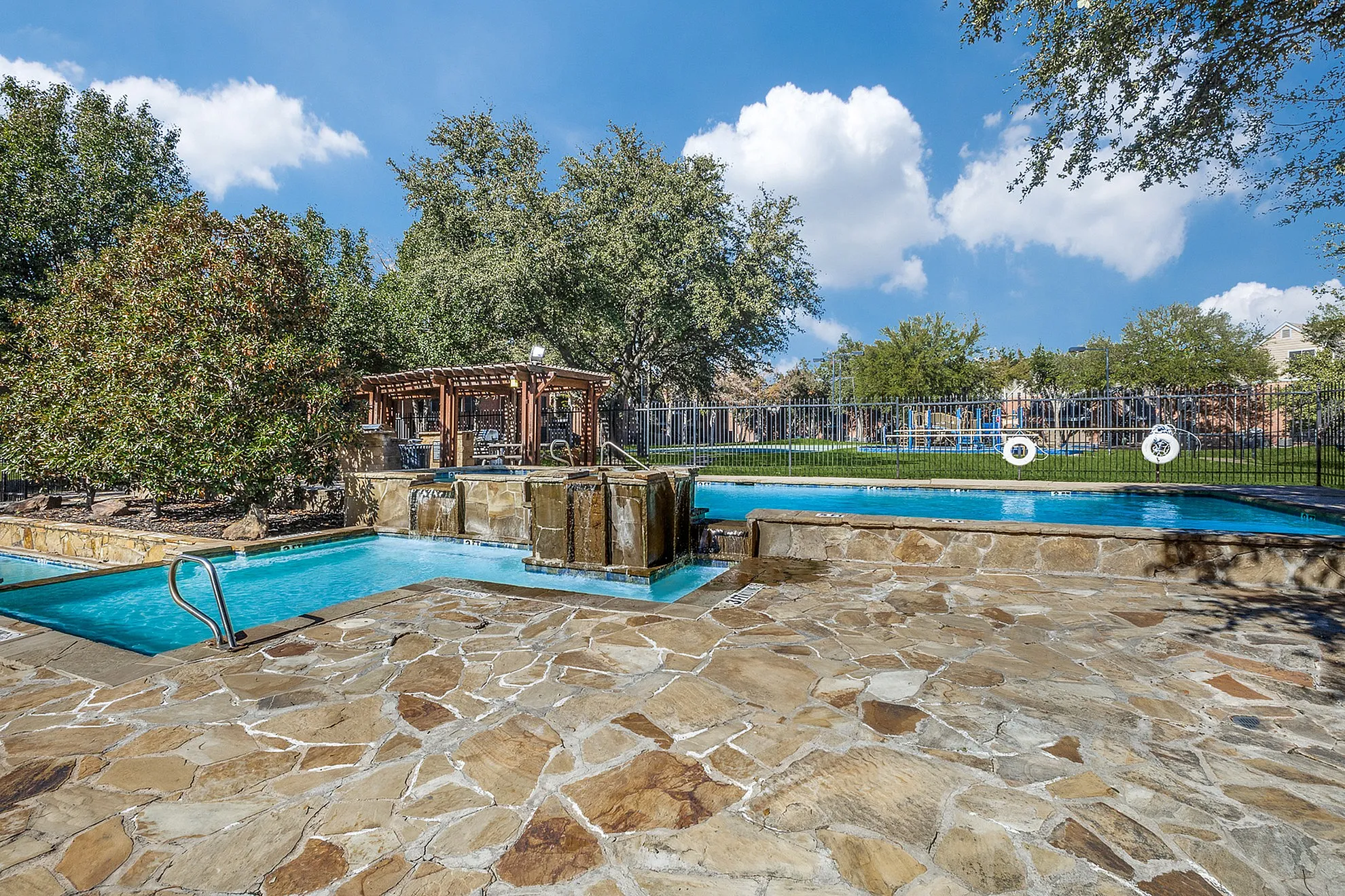 Luxurious outdoor pool area with cascading waterfalls, lush greenery, and a cozy pergola, perfect for relaxation and leisure.
