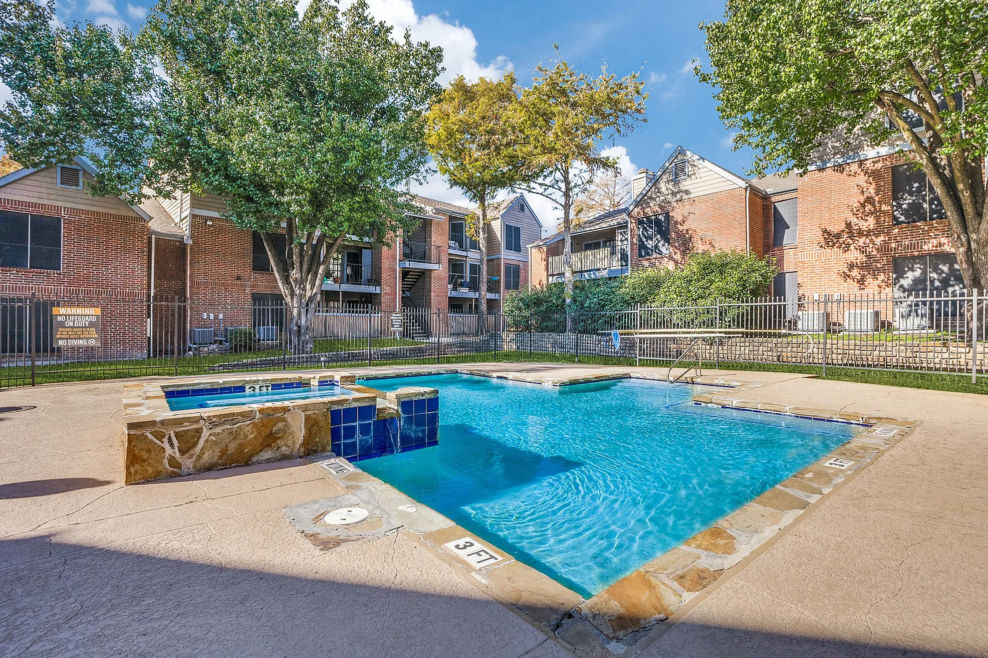 Inviting outdoor pool area amidst lush greenery and charming brick apartment buildings, perfect for relaxation.