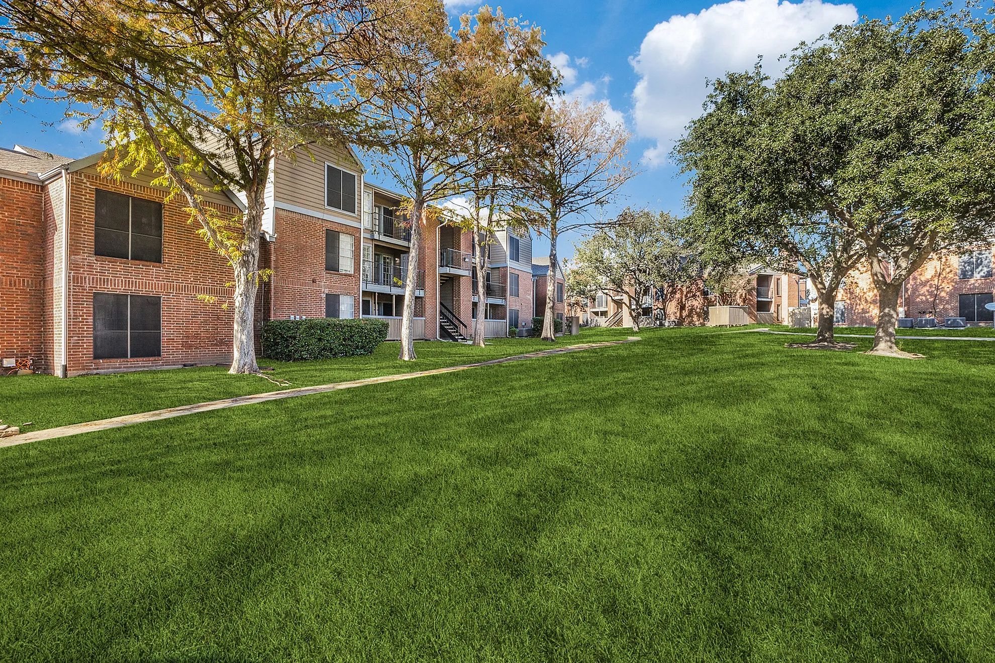 Inviting apartment complex with lush greenery, vibrant trees, and modern brick exteriors under a bright blue sky.