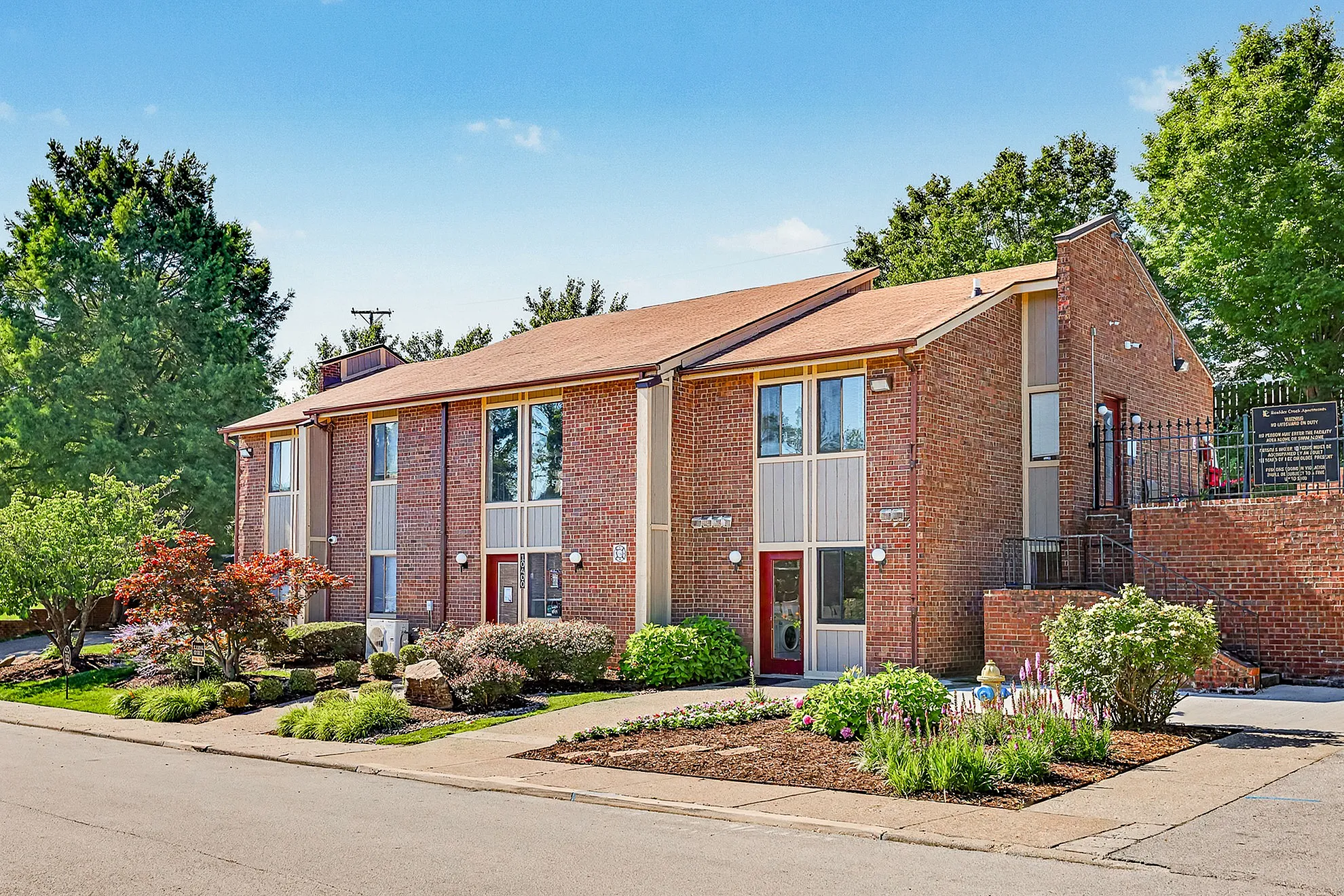 Charming brick apartment building with large windows surrounded by lush landscaping and vibrant flower beds in a serene residential setting.