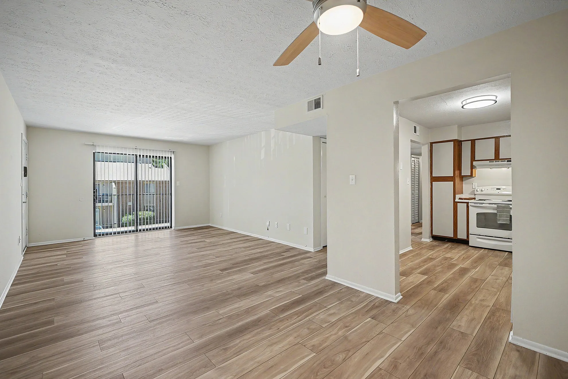 Spacious living area with modern wood flooring, ceiling fan, and large sliding glass doors leading to a bright balcony; adjacent kitchen with stylish cabinetry and contemporary white appliances.
