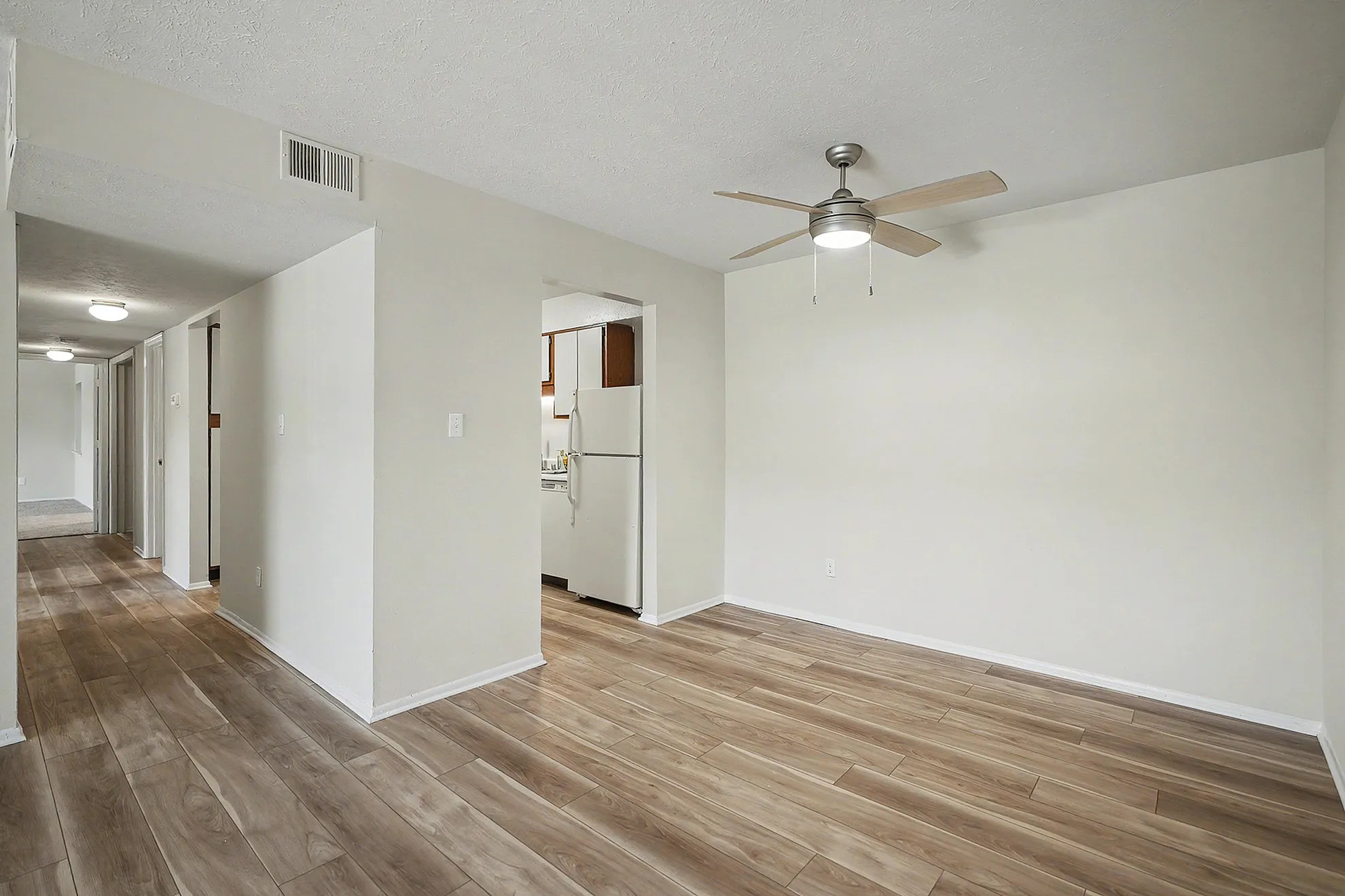 Bright apartment interior with modern wooden flooring, ceiling fan, and a glimpse into a cozy kitchen area.