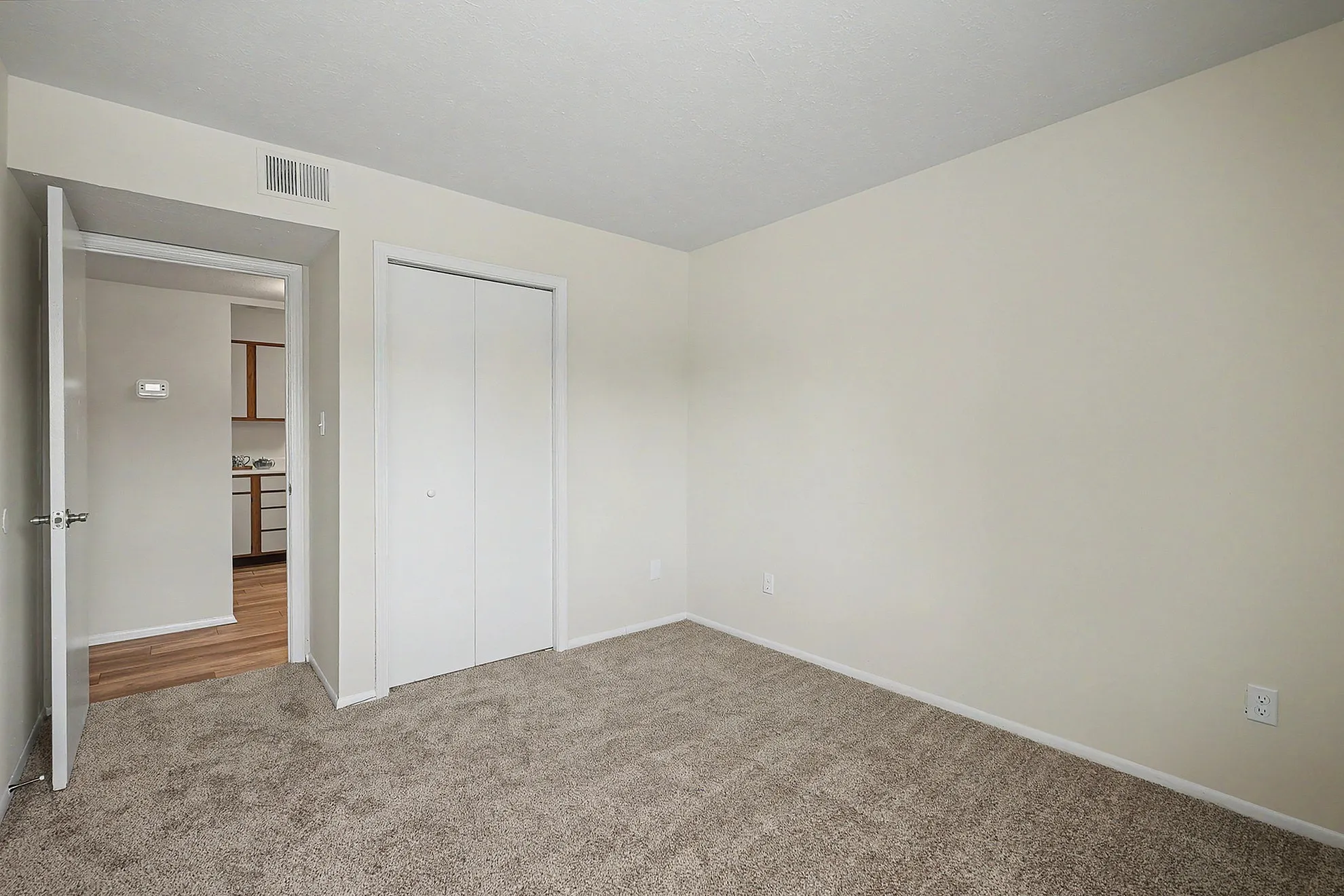Bright, empty room with beige carpet, white walls, and a cozy atmosphere connecting to a charming kitchen.