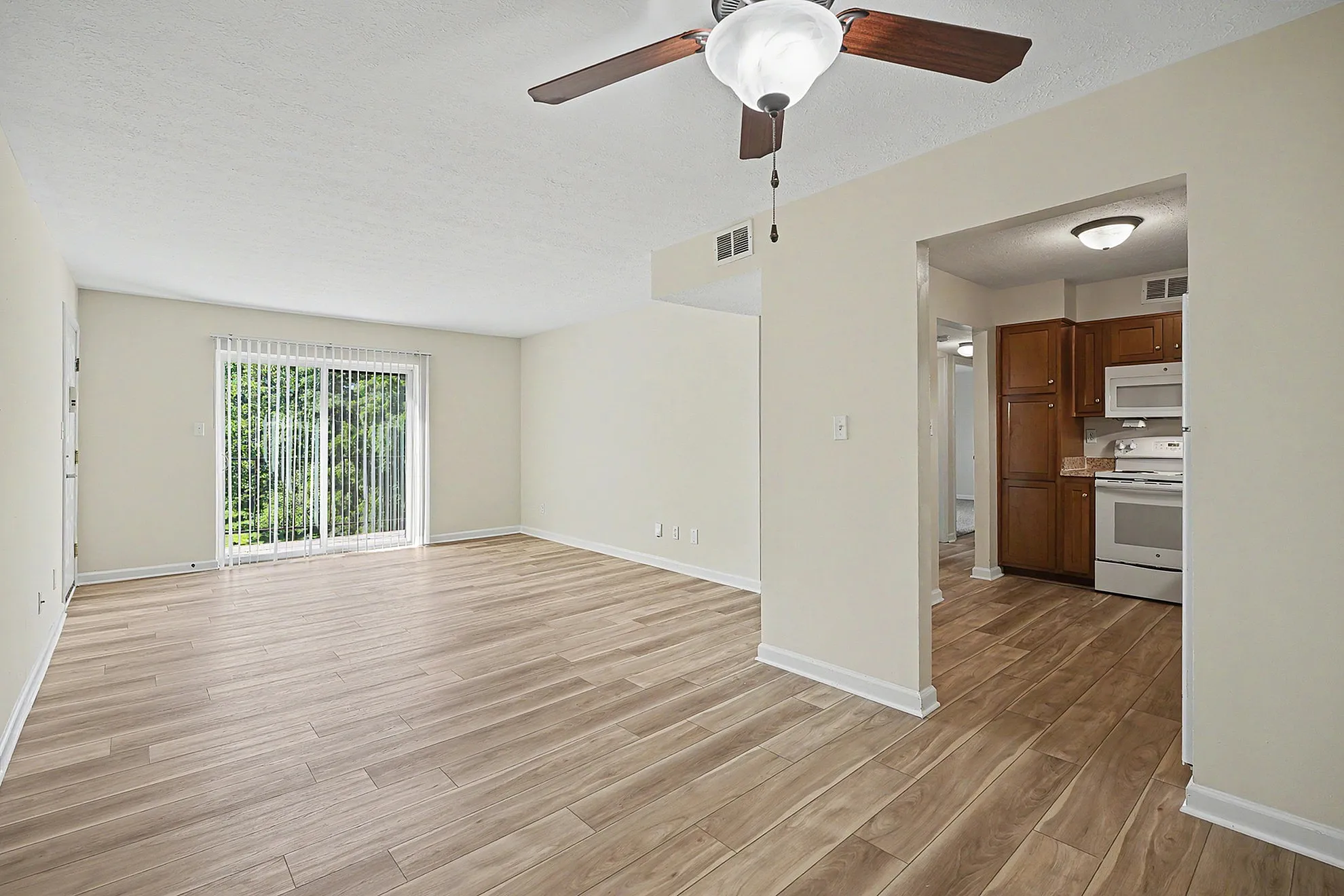 Bright and spacious living area with elegant wooden flooring, modern ceiling fan, and a cozy kitchen opening featuring classic wood cabinetry. Stunning natural light pours in through large sliding glass doors, offering a serene view of the greenery outside.