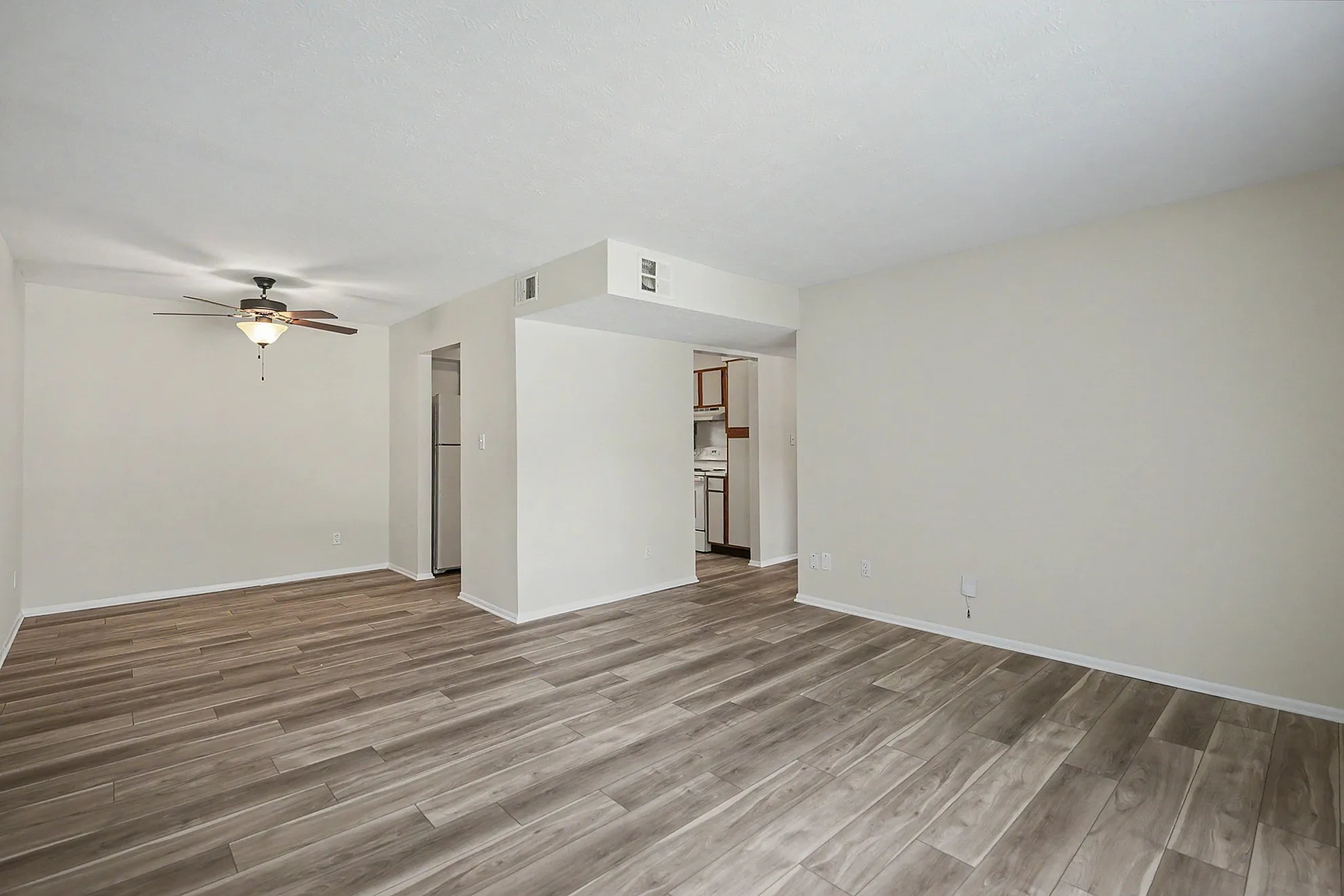 Modern apartment living room with elegant wood-style flooring and a ceiling fan, opening into a cozy kitchen area.