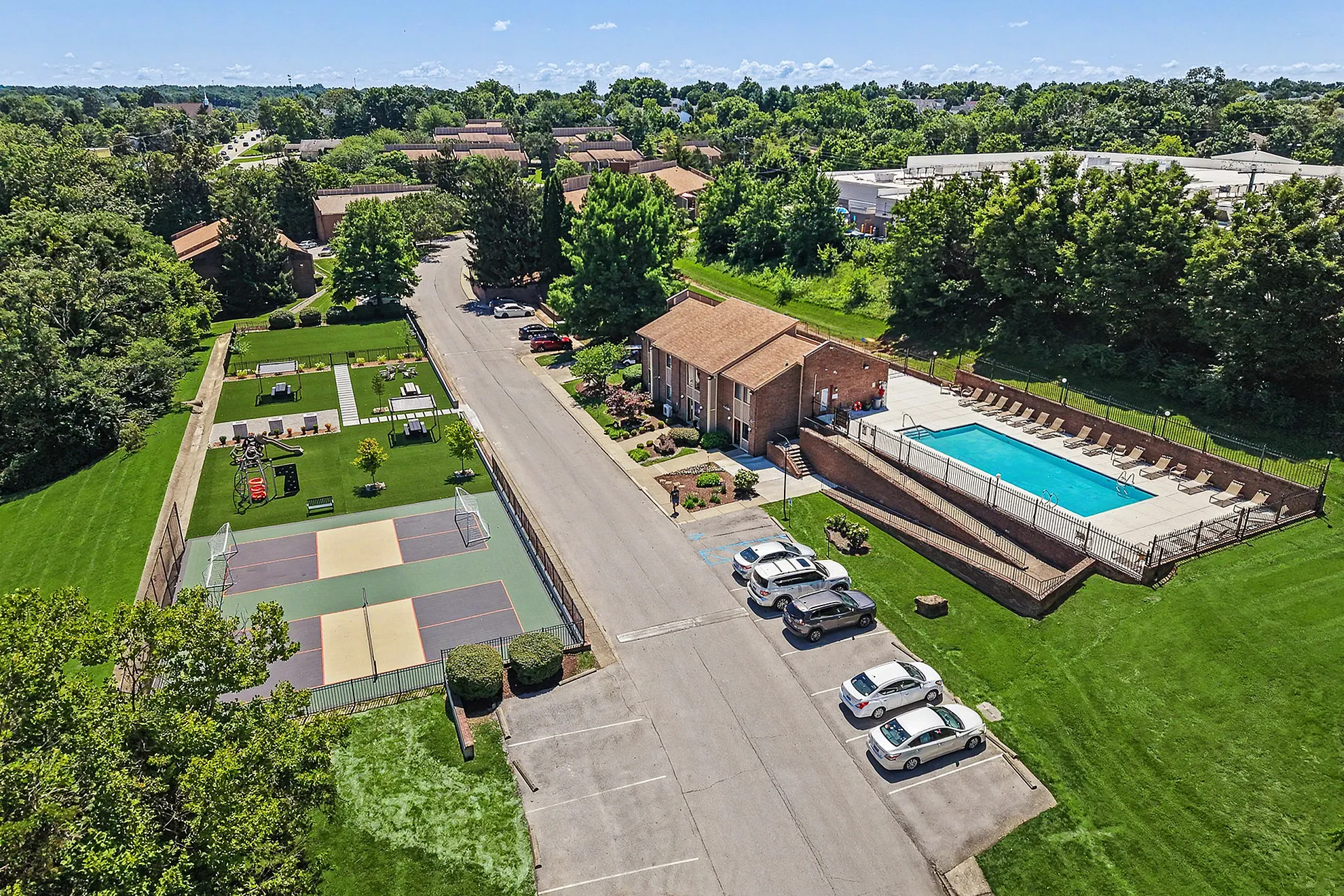 Aerial view of a vibrant community with a tennis court, lush green areas, a modern swimming pool, and ample parking. Ideal for active lifestyles.