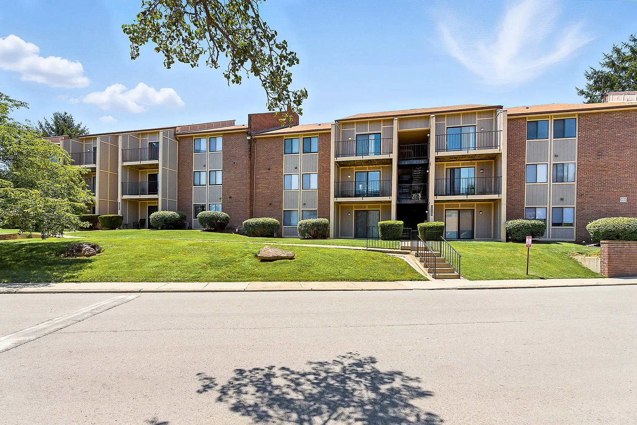 Charming brick apartment complex with spacious balconies, surrounded by lush greenery and bright blue skies.