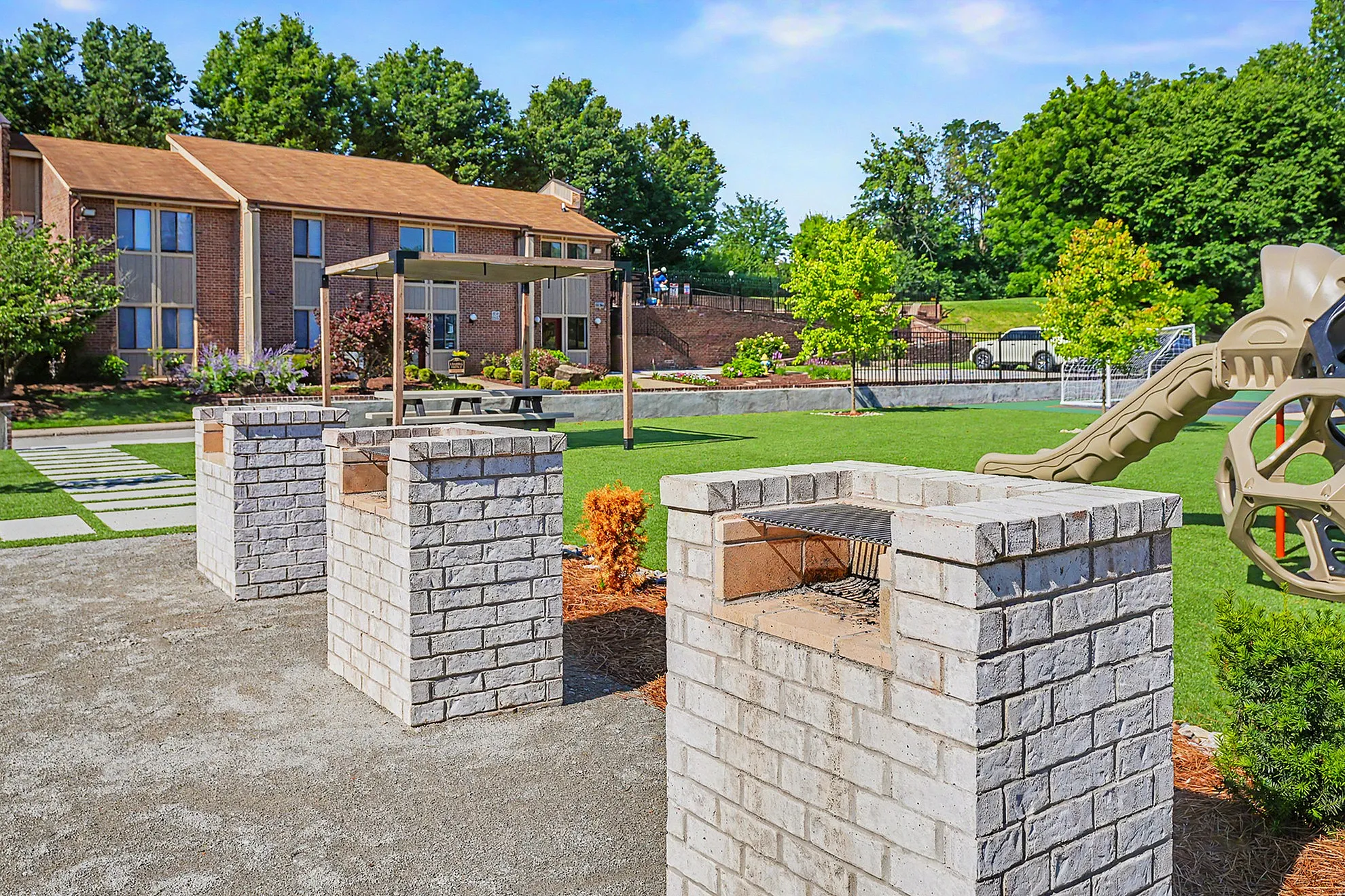 Charming outdoor communal area with brick barbecue pits, lush greenery, and a modern playground in a welcoming apartment community.