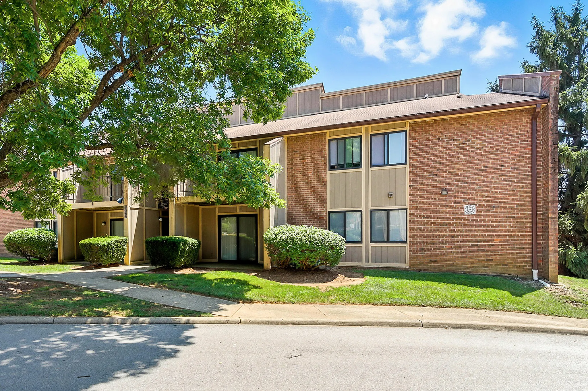 Charming brick apartment building surrounded by lush greenery and vibrant landscaping, featuring modern windows and a welcoming entrance.