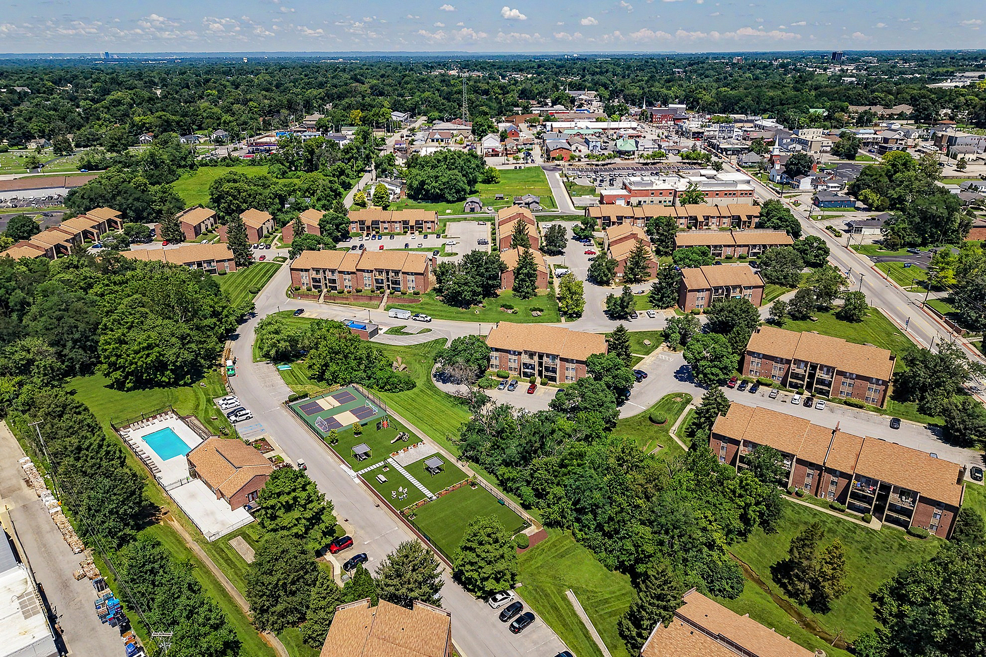 Aerial view of a vibrant apartment community featuring multiple brick buildings, lush greenery, a sparkling pool, and recreational spaces in a serene neighborhood.