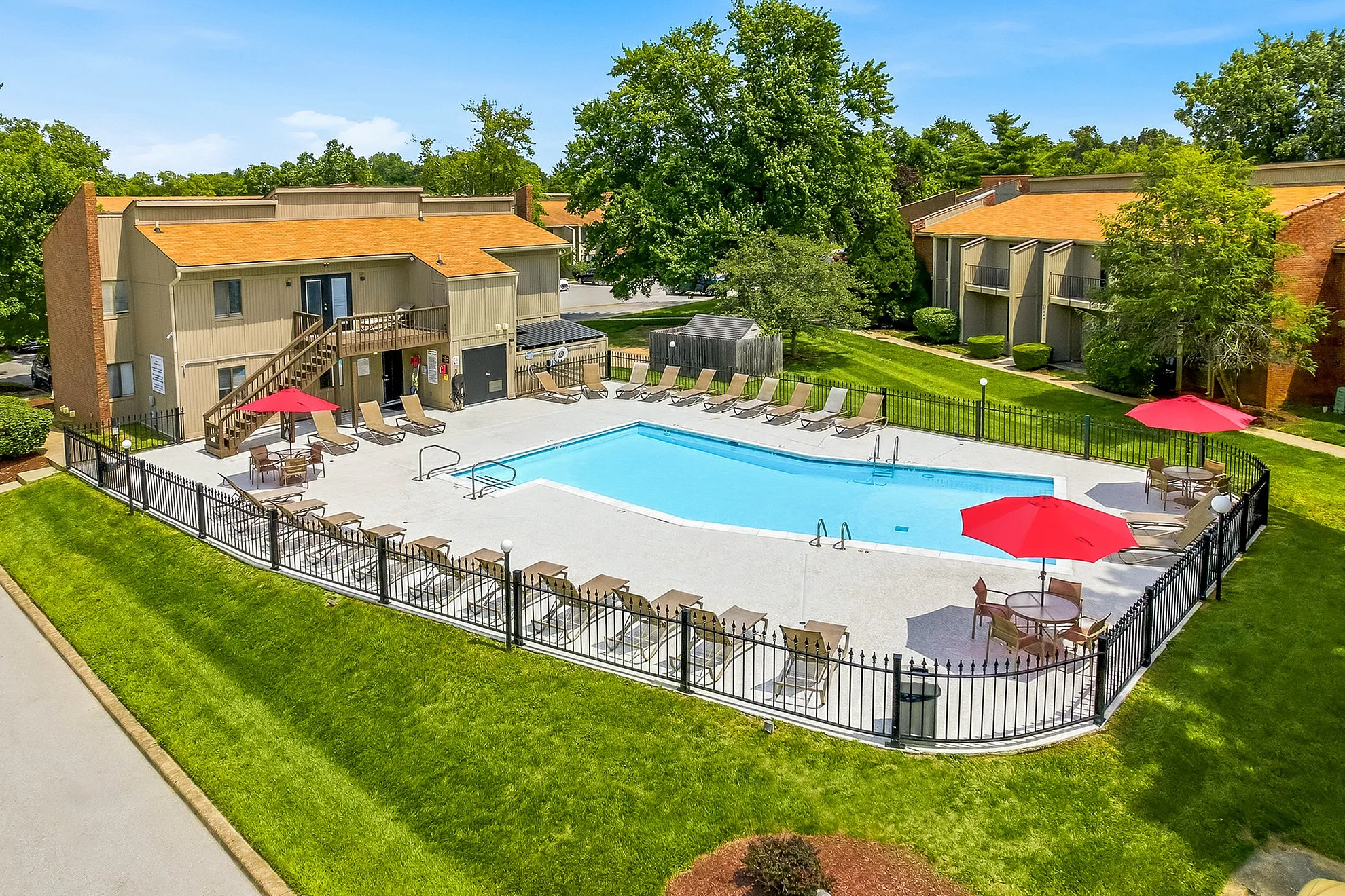 Inviting apartment complex featuring a sparkling outdoor pool with lounge chairs, surrounded by lush greenery and red umbrellas on a sunny day.