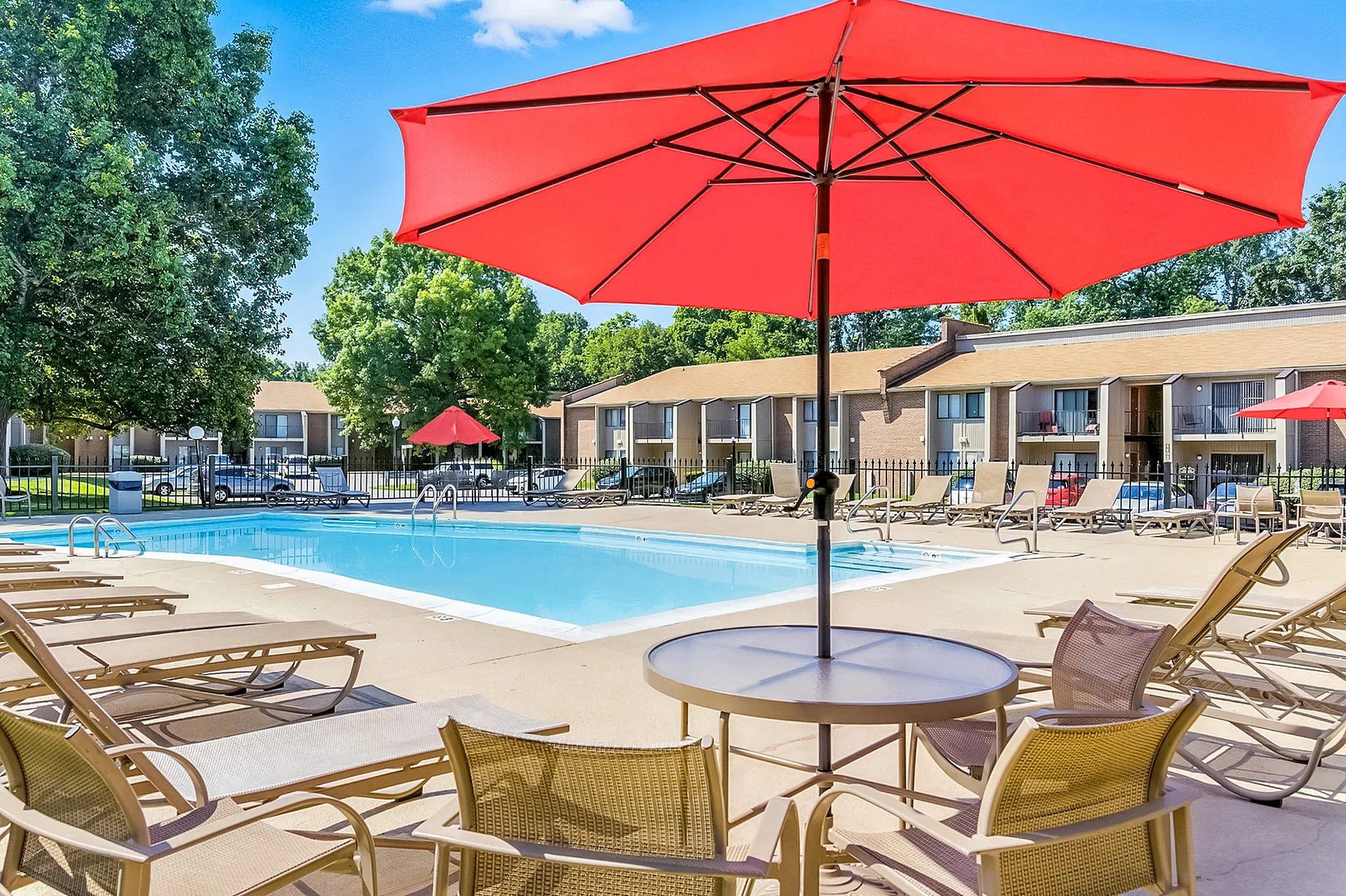 Elegant apartment complex with a pristine swimming pool surrounded by stylish lounge chairs and vibrant red umbrellas, perfect for relaxing weekends.