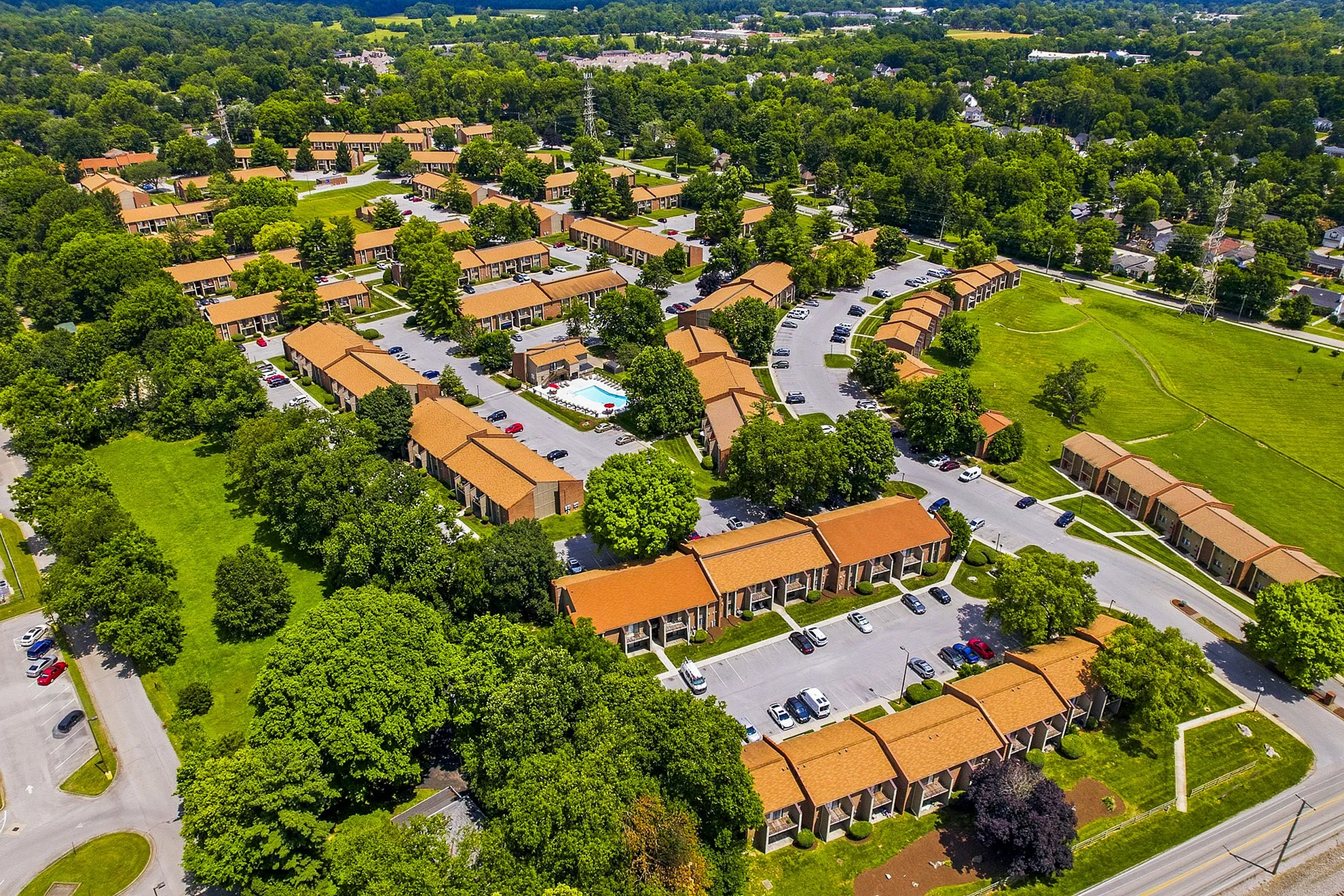 Vibrant aerial view of a beautifully landscaped apartment complex with terracotta roofs surrounded by lush greenery, featuring ample parking and a refreshing pool area.