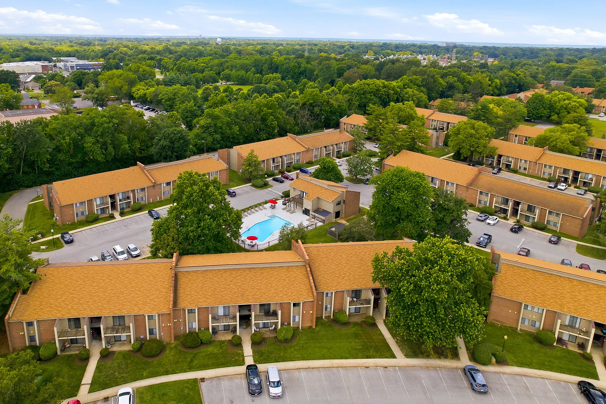 Aerial view of a beautifully landscaped apartment complex with charming brown roofs, lush greenery, and a sparkling blue swimming pool at the center.
