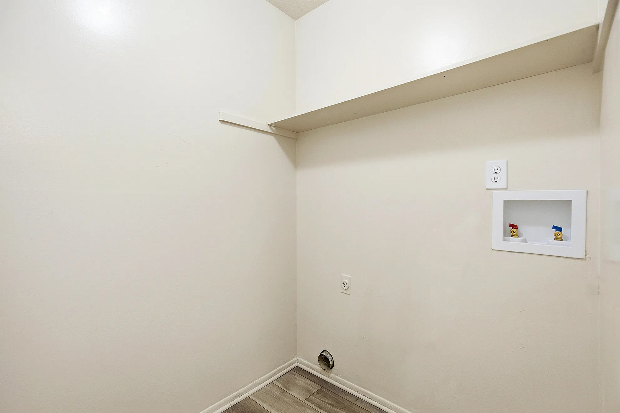 Spacious laundry area with sleek white walls, convenient wall shelf, and utility plug, ready for modern appliances.