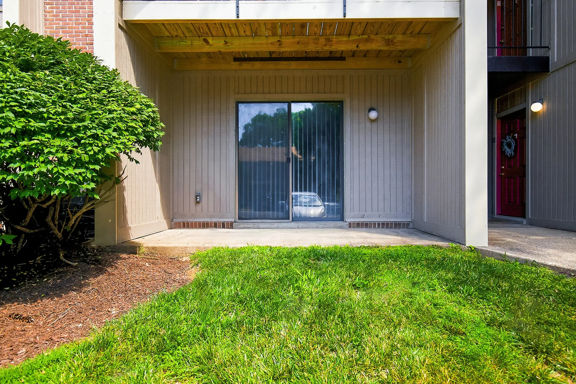 Ground-level apartment patio with sliding glass doors, lush greenery, and modern design.