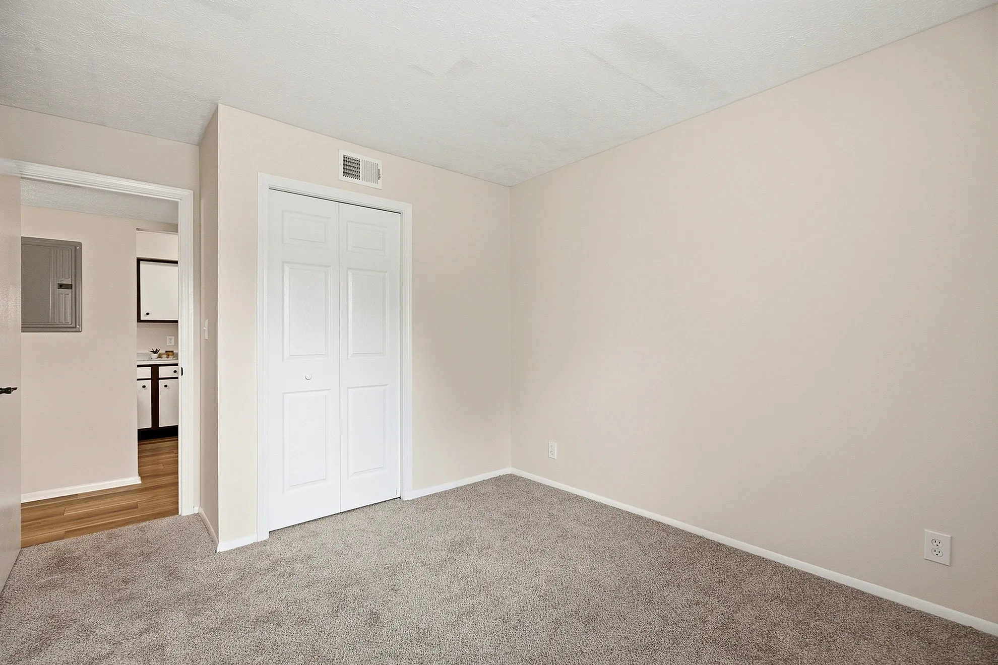 Bright, carpeted bedroom with cream walls and a spacious closet, offering a glimpse into the stylish kitchen with wood flooring.