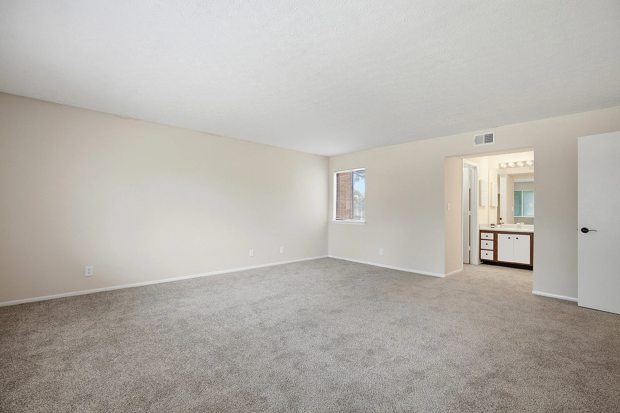 Bright and expansive bedroom with soft carpet flooring, neutral walls, and a window for natural light, leading to a private ensuite bathroom.