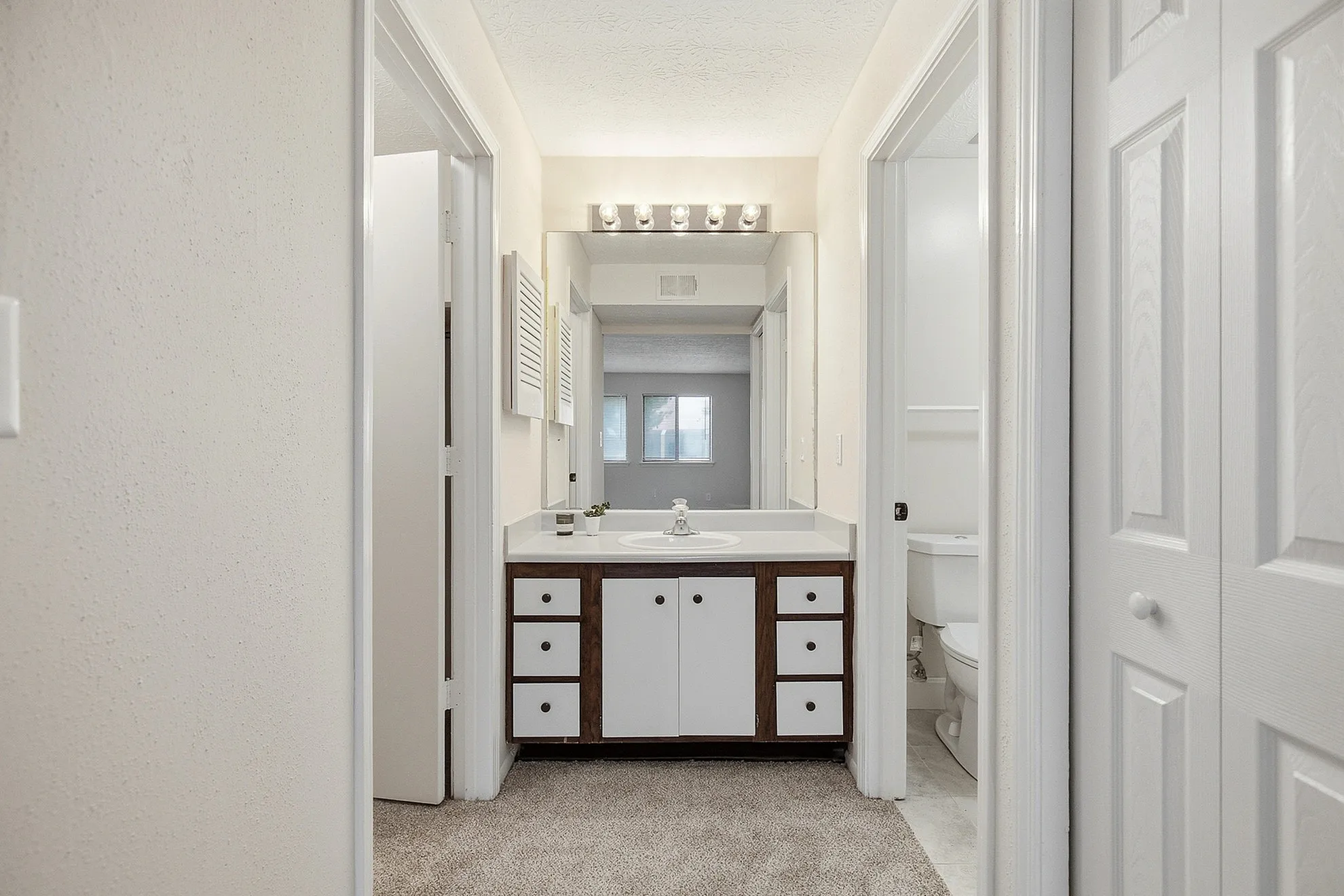 Modern apartment bathroom featuring a sleek vanity with ample storage, bright lighting, and a plush carpeted hallway.