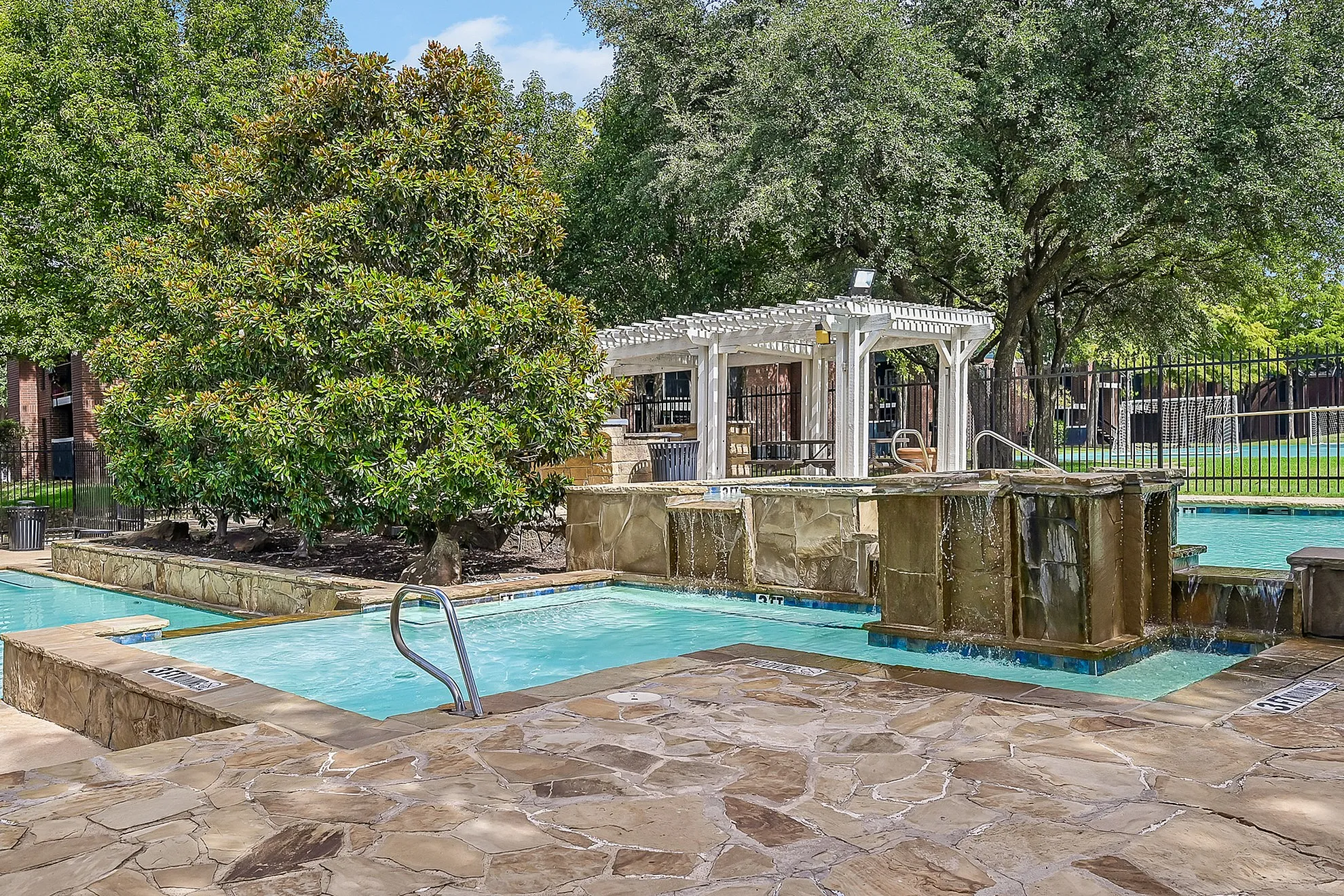 Luxurious outdoor pool area with cascading waterfalls, surrounded by lush greenery and a charming pergola for relaxation.