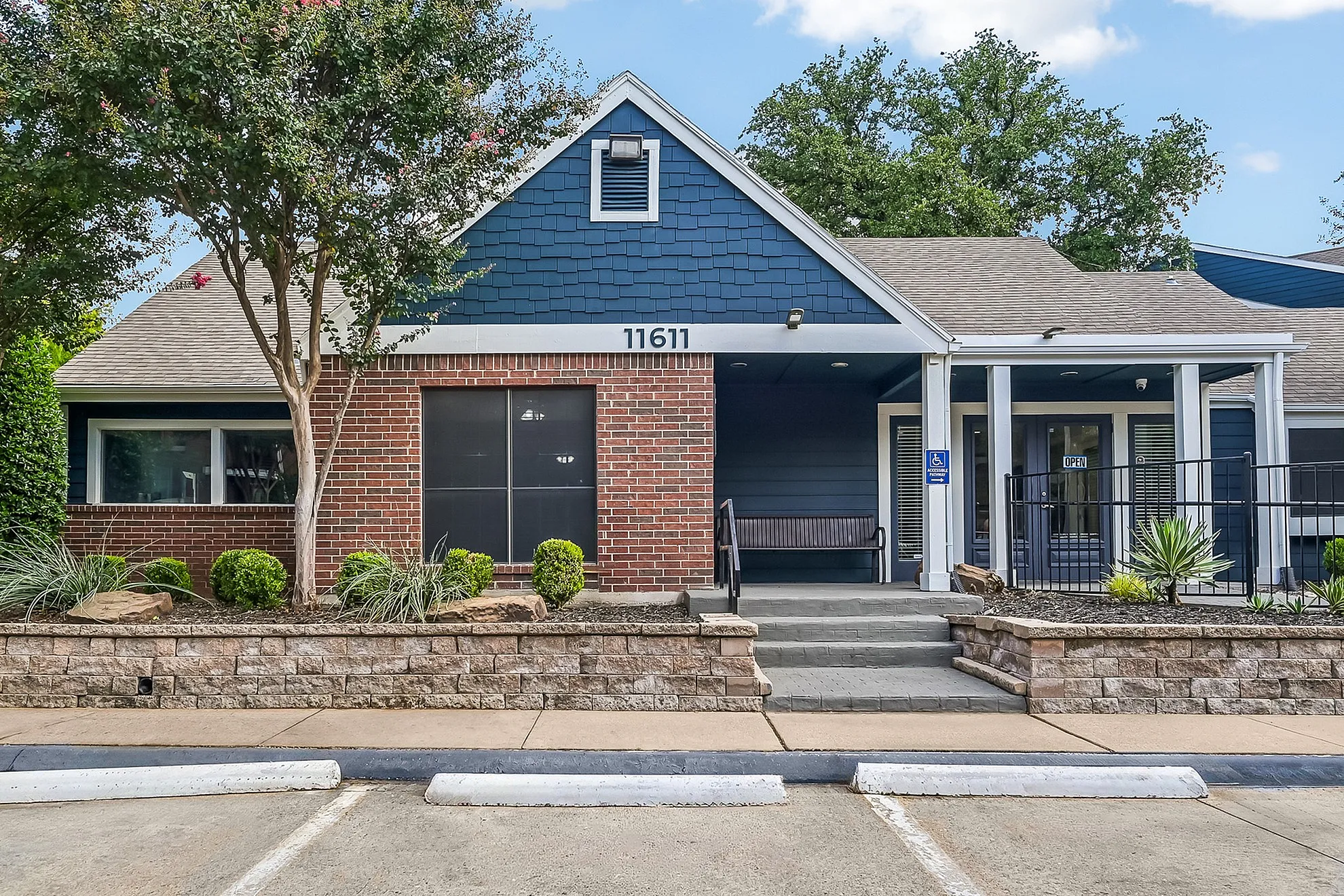 Charming brick and blue-paneled apartment entrance with accessible features, surrounded by lush greenery and modern landscaping.